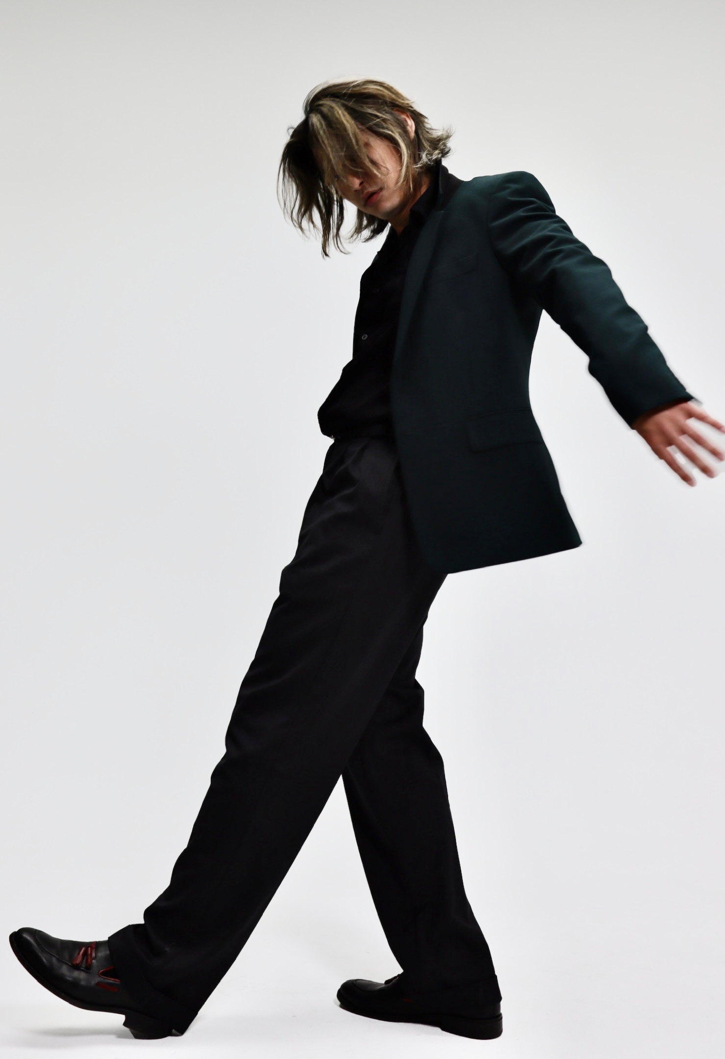 A person with long hair dressed in a black suit, black shirt, and black shoes, in a dynamic pose against a plain white background.