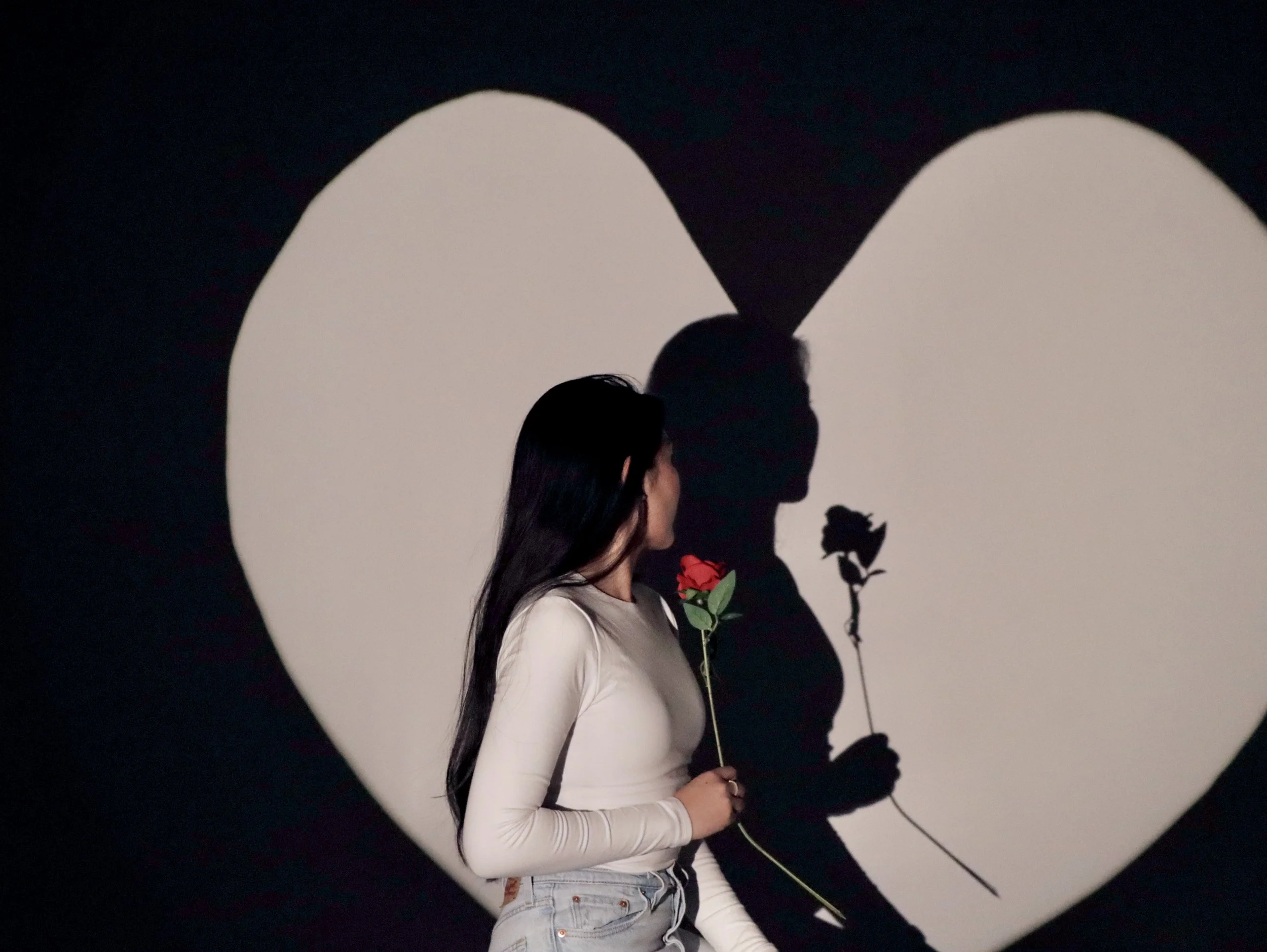 A woman is holding a red rose and looking at her shadow behind a heart-shaped projection on the wall, creating a romantic scene.