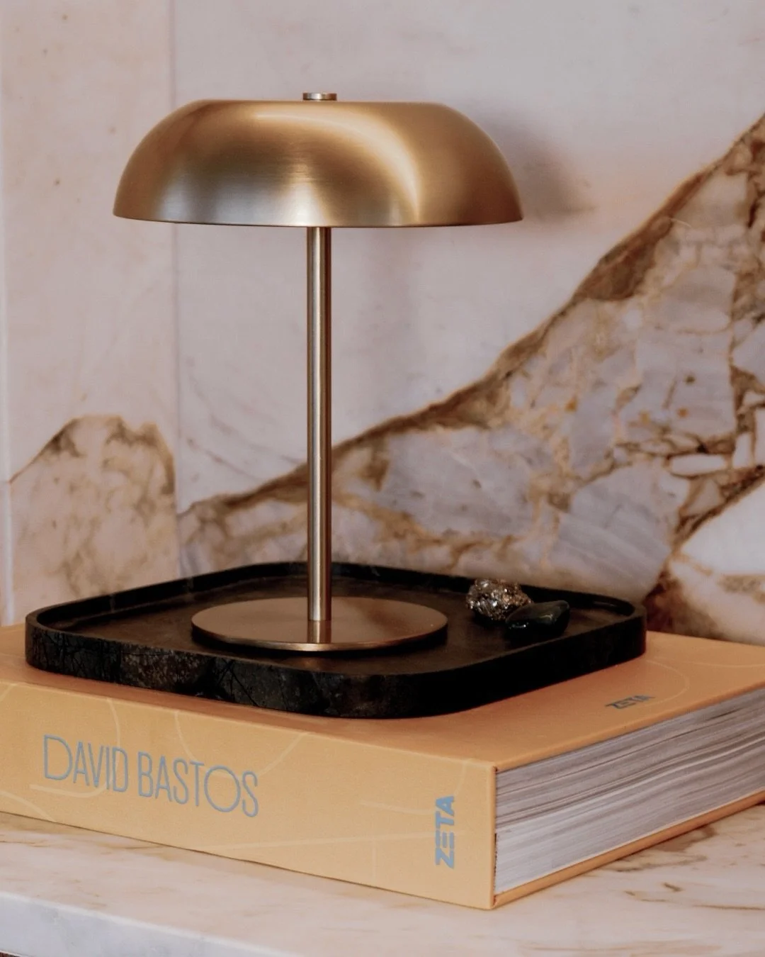 Luz de mesa dourada com base circular, colocada sobre um livro intitulado "David Bastos" na mesa ao lado de pedras decorativas, com fundo de parede de mármore.