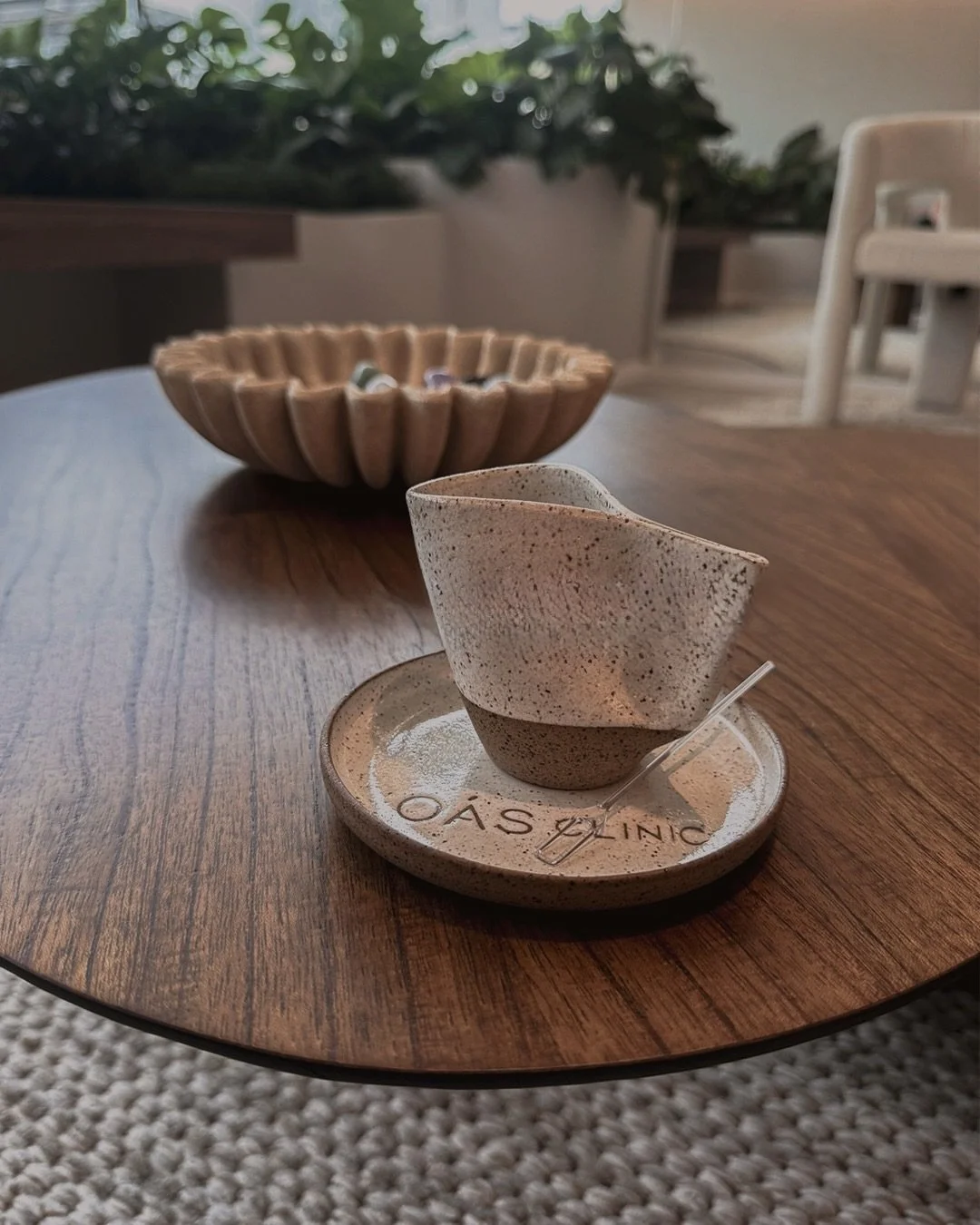 Xícara de cerâmica com pluma de chá, em um suporte com a inscrição 'OASIS CLINIC' sobre uma mesa de madeira, com uma tigela de madeira ao fundo e sofá branco ao fundo.