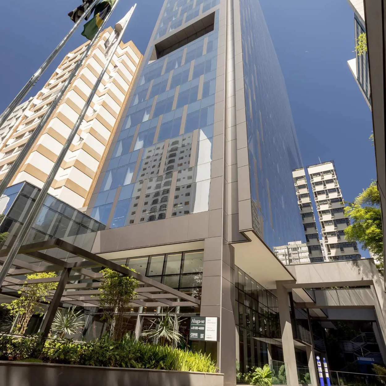 Prédio de escritórios com fachada de vidro refletindo outros edifícios e céu azul, com áreas de plantas na frente e bandeiras ao lado.