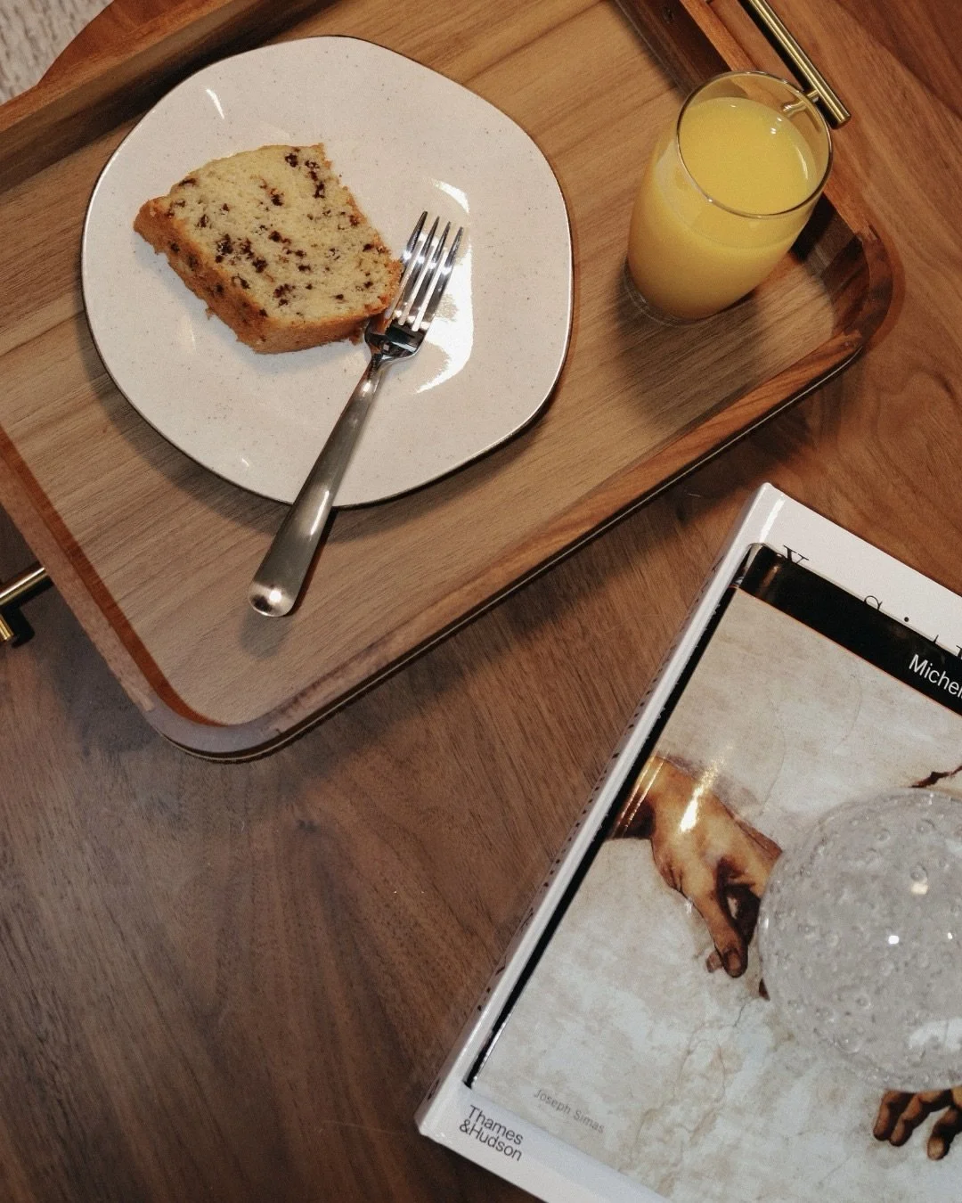 Porção de bolo de chocolate em um prato branco, garfo ao lado, copo de suco de laranja e livro sobre uma bandeja de madeira sobre uma mesa de madeira.