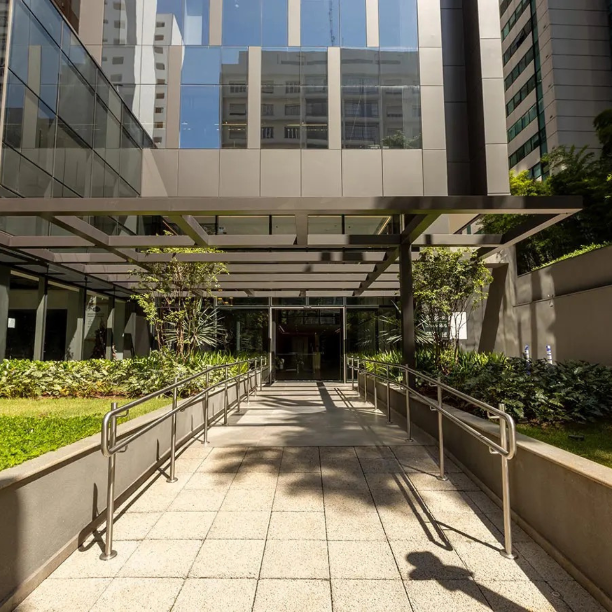 Entrada de edifício moderno com acessibilidade, plantas e guarda-corpos em metal, fachada de vidro refletindo outros prédios.