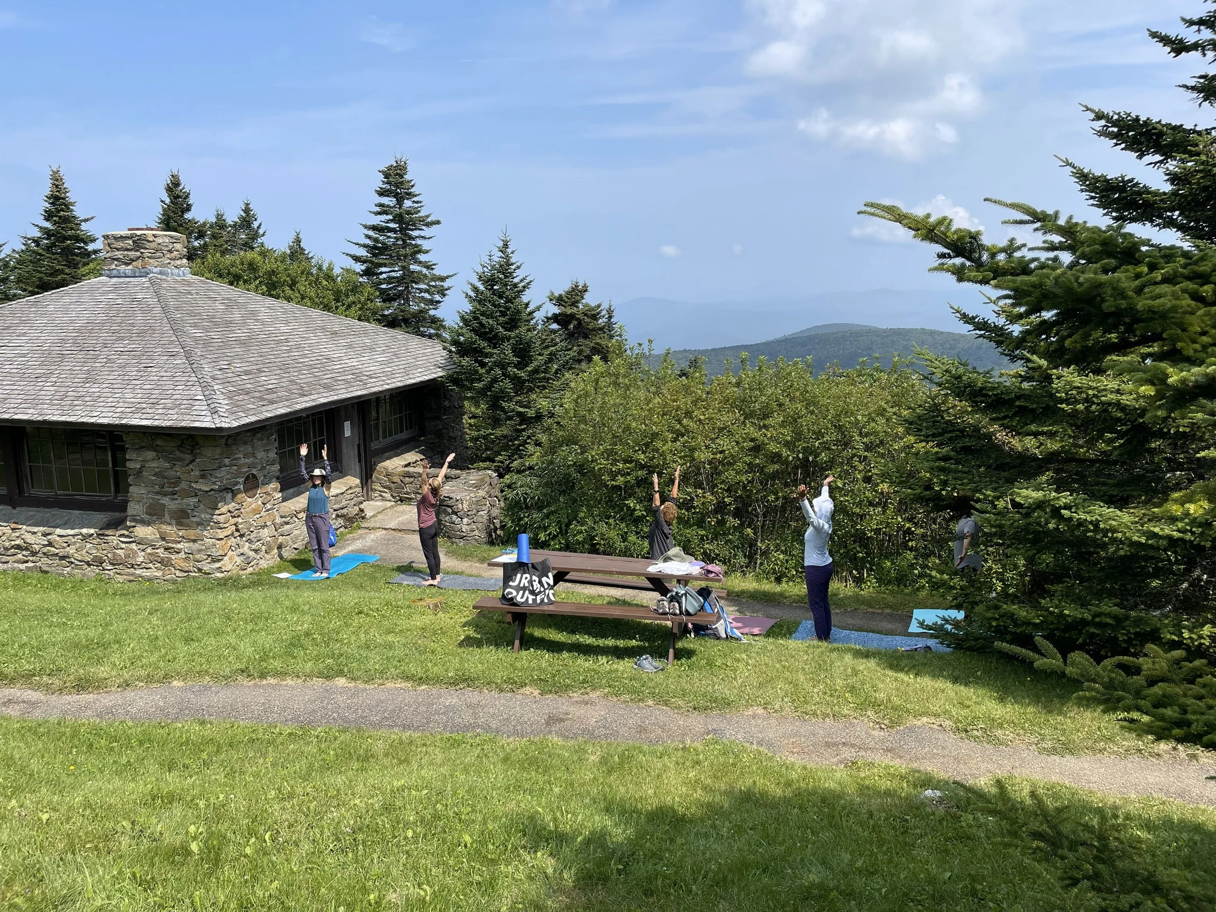 People practicing yoga outdoors on a grassy hill near a stone house with a mountain view in the background.