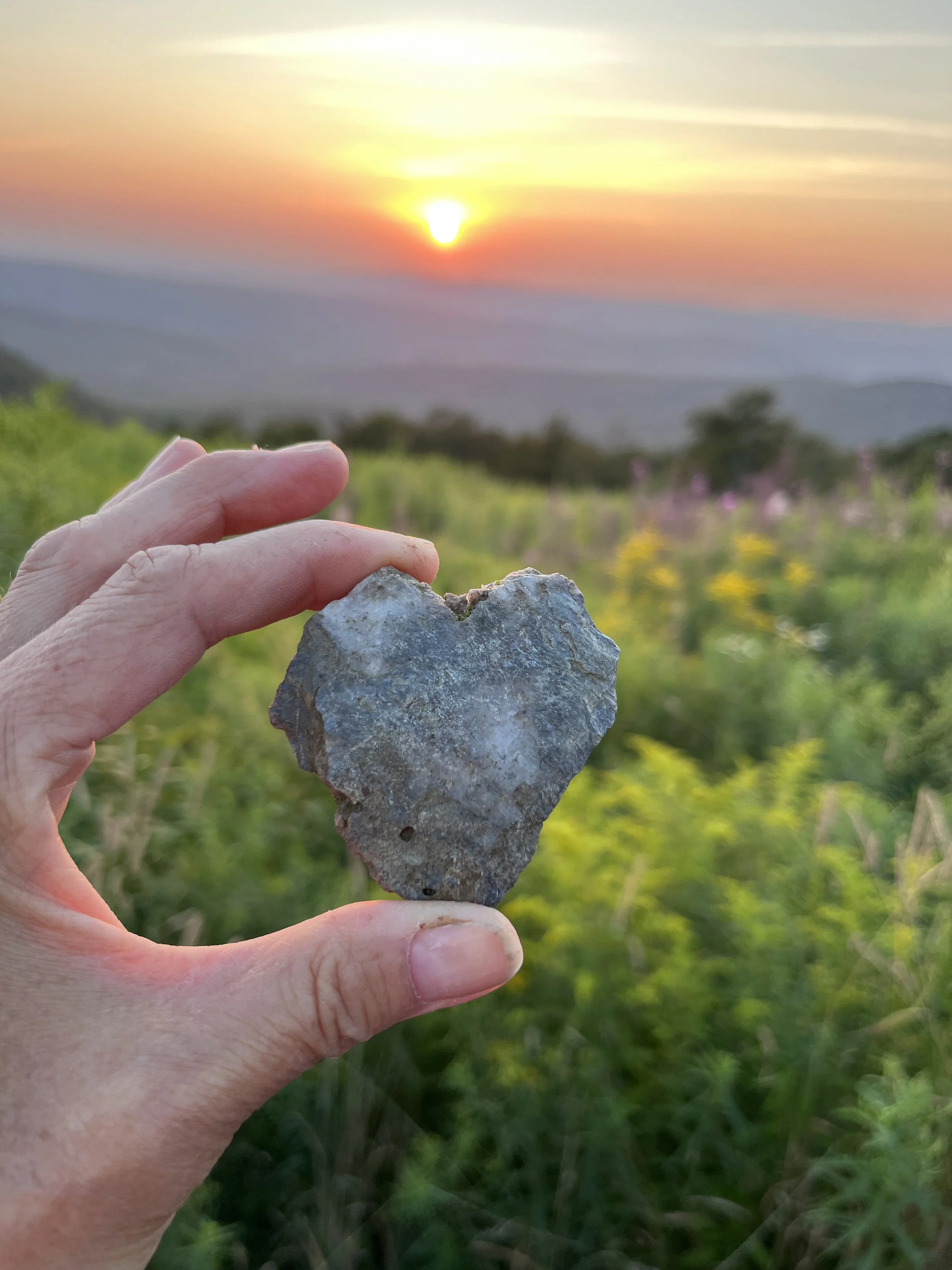 A person's hand holding a heart-shaped stone in the foreground, with a scenic landscape of hills, green fields, and a sunset in the background.