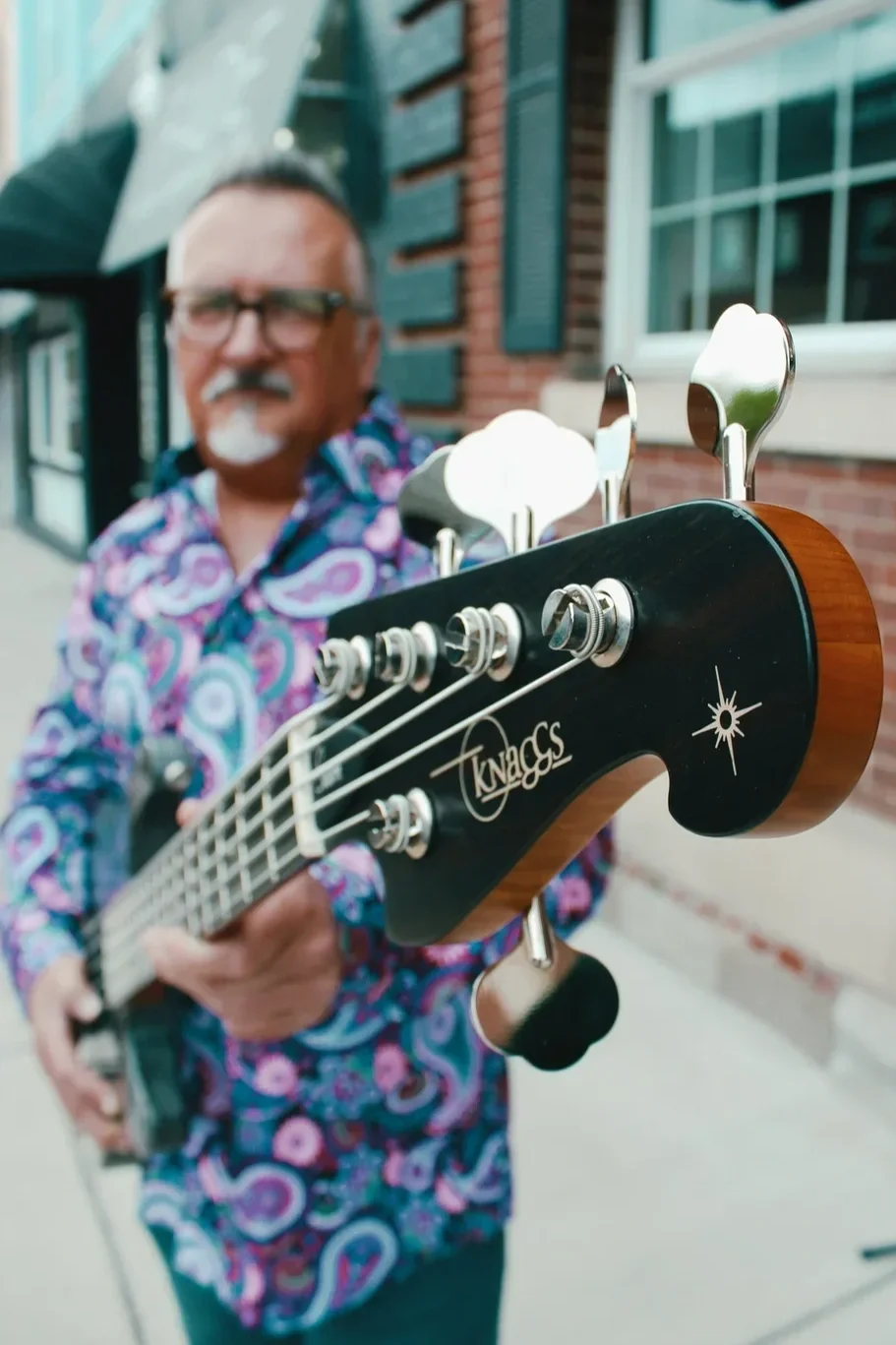 Close-up of a black Knaggs bass guitar headstock , held by a man with glasses and a colorful, psychedelic-patterned shirt, standing outside a brick building.