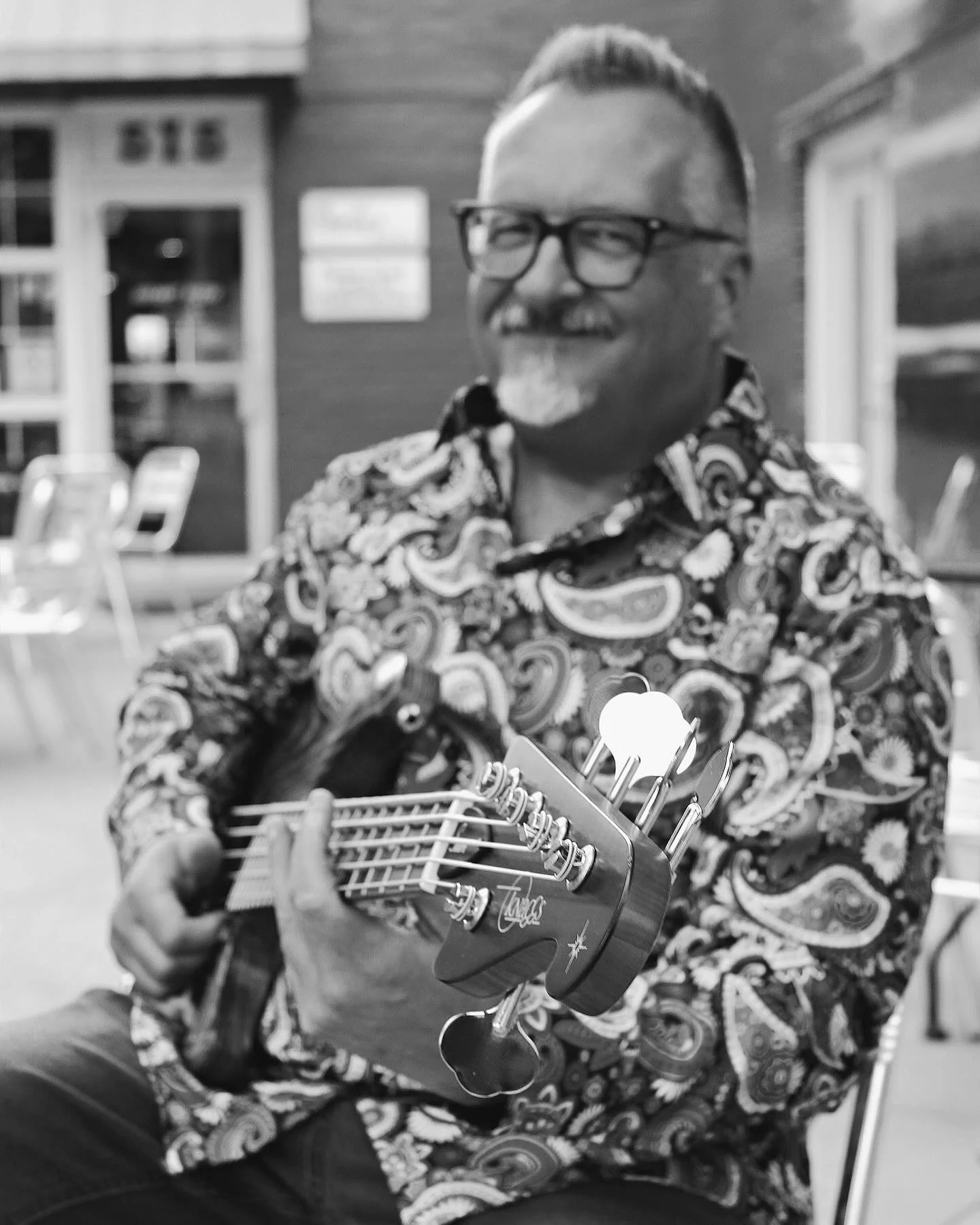 Man with glasses, playing a Knaggs bass guitar with a patterned shirt in a casual outdoor setting.