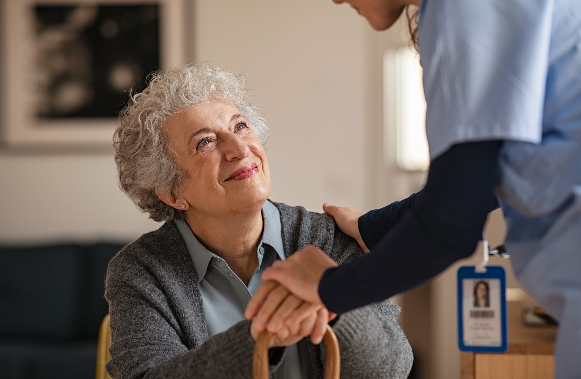 An elderly woman with gray, curly hair smiling and looking up at a caregiver, who is gently holding her hands, in a supportive and caring manner.