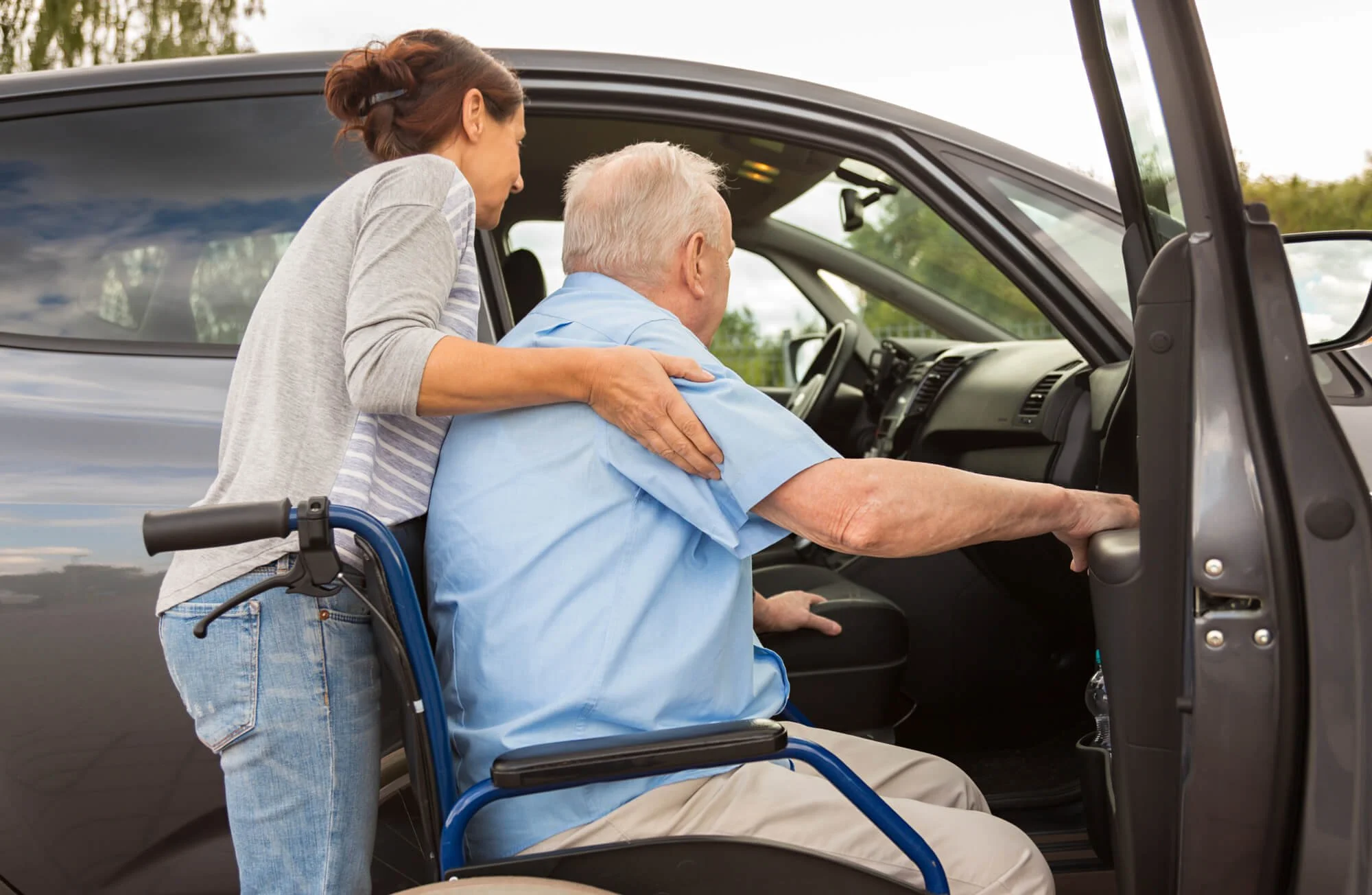 An elderly man in a wheelchair getting assistance from a woman as he prepares to get into a vehicle. Concierge home care Houston — Care Guild Houston caregiver,