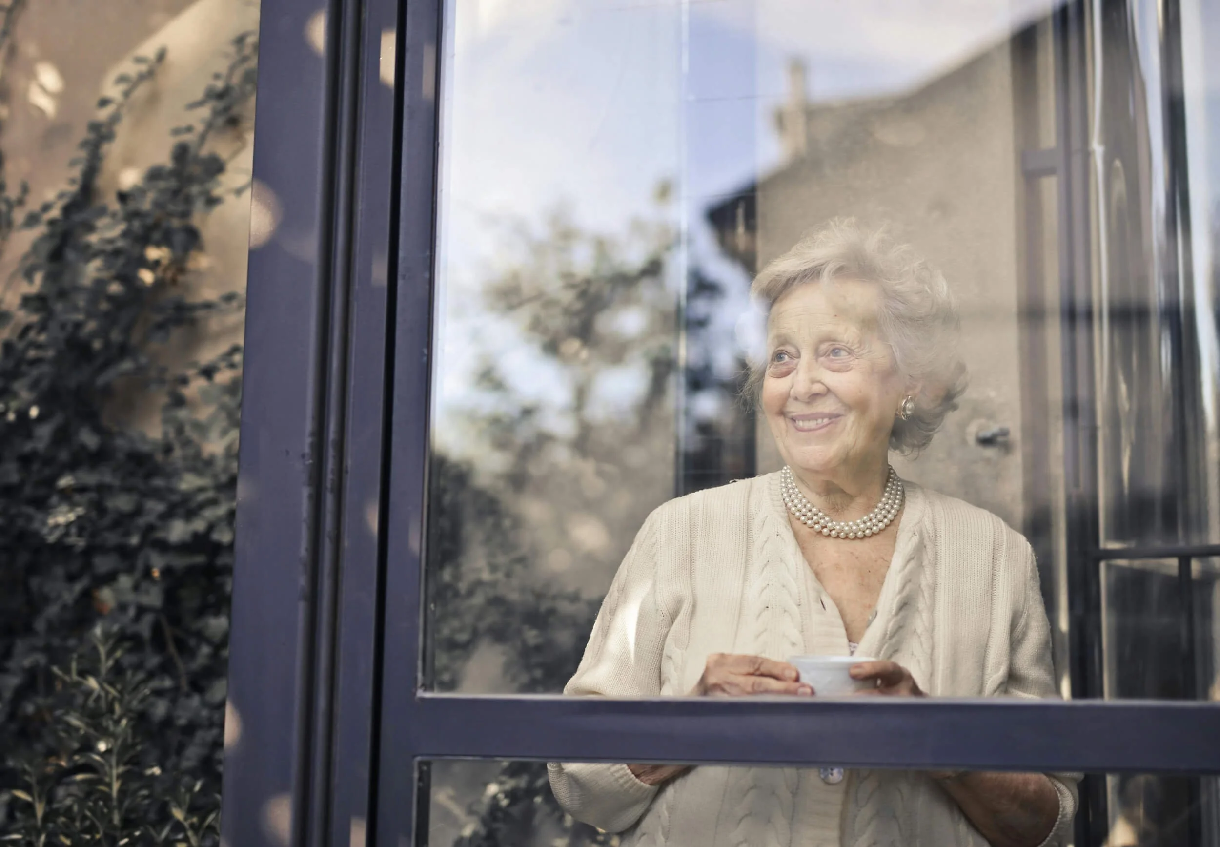 An elderly woman smiling while drinking tea behind a glass door, with trees and house in the background. In-home dementia care support Houston TX
