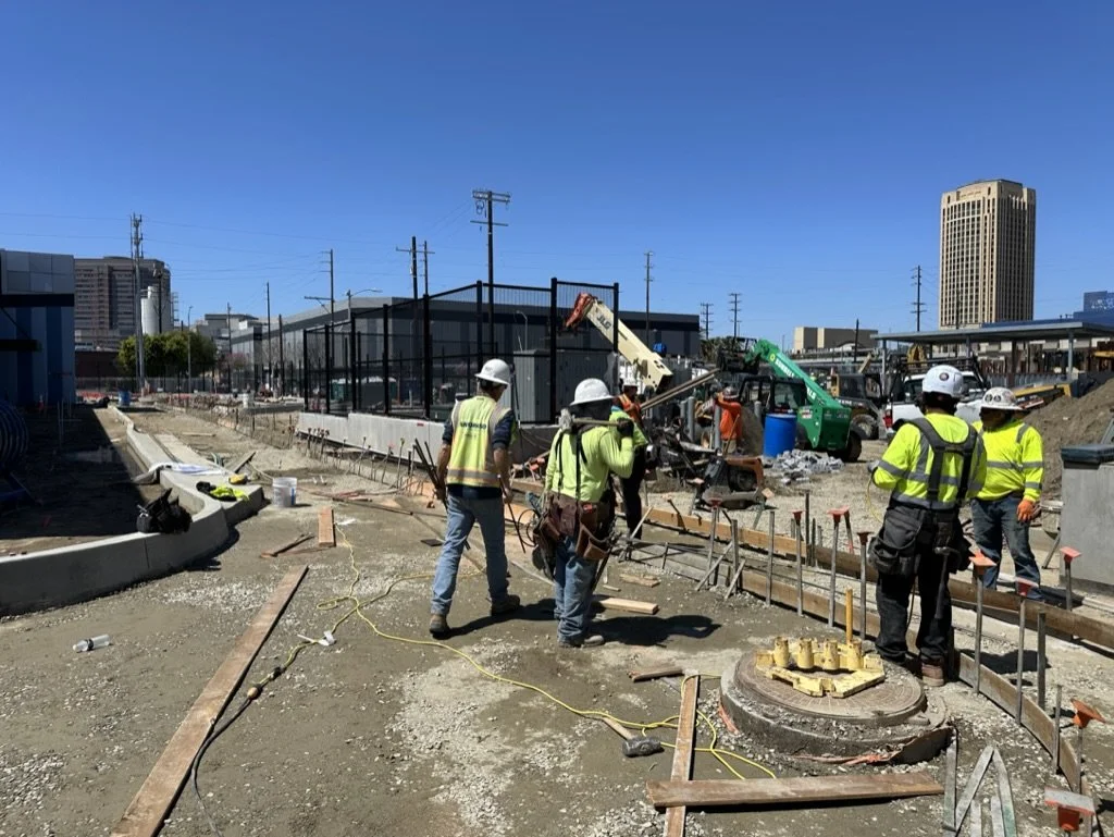 Construction workers in safety vests and helmets working on a building site with machinery, tools, and construction materials under a clear blue sky.