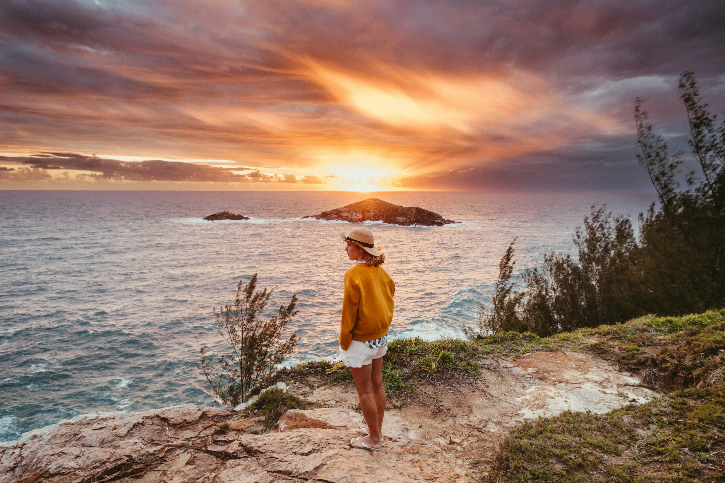 A woman standing on a rocky ledge over looking a bright yellow sunset over the ocean. The sky is cloudy and the waves are peaceful. She's wearing an orange sweatshirt, white shorts, and a straw hat.