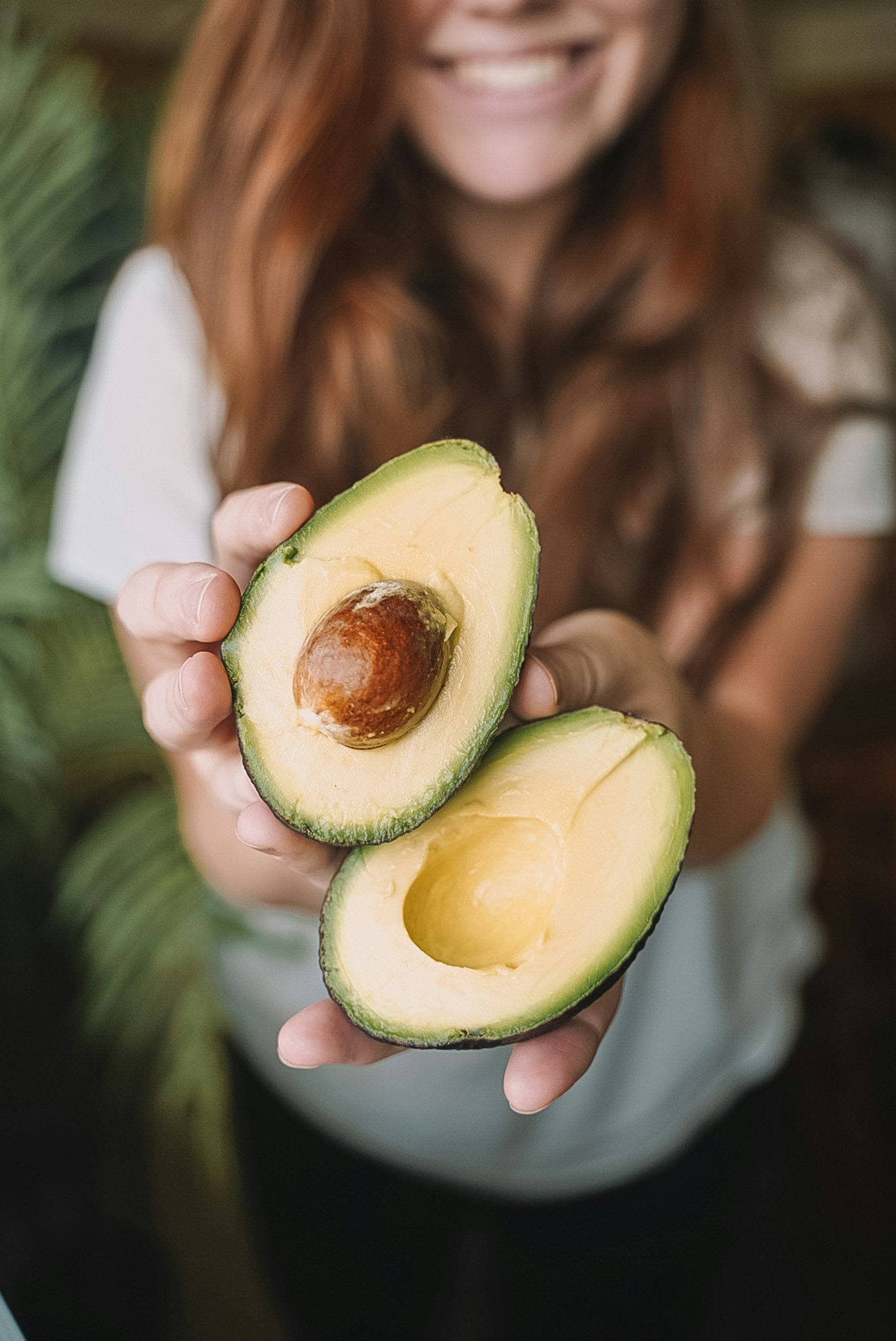 Woman holding an avocado. The avocado is cut in half. She is showing the side of the fruit. You cannot see her face.