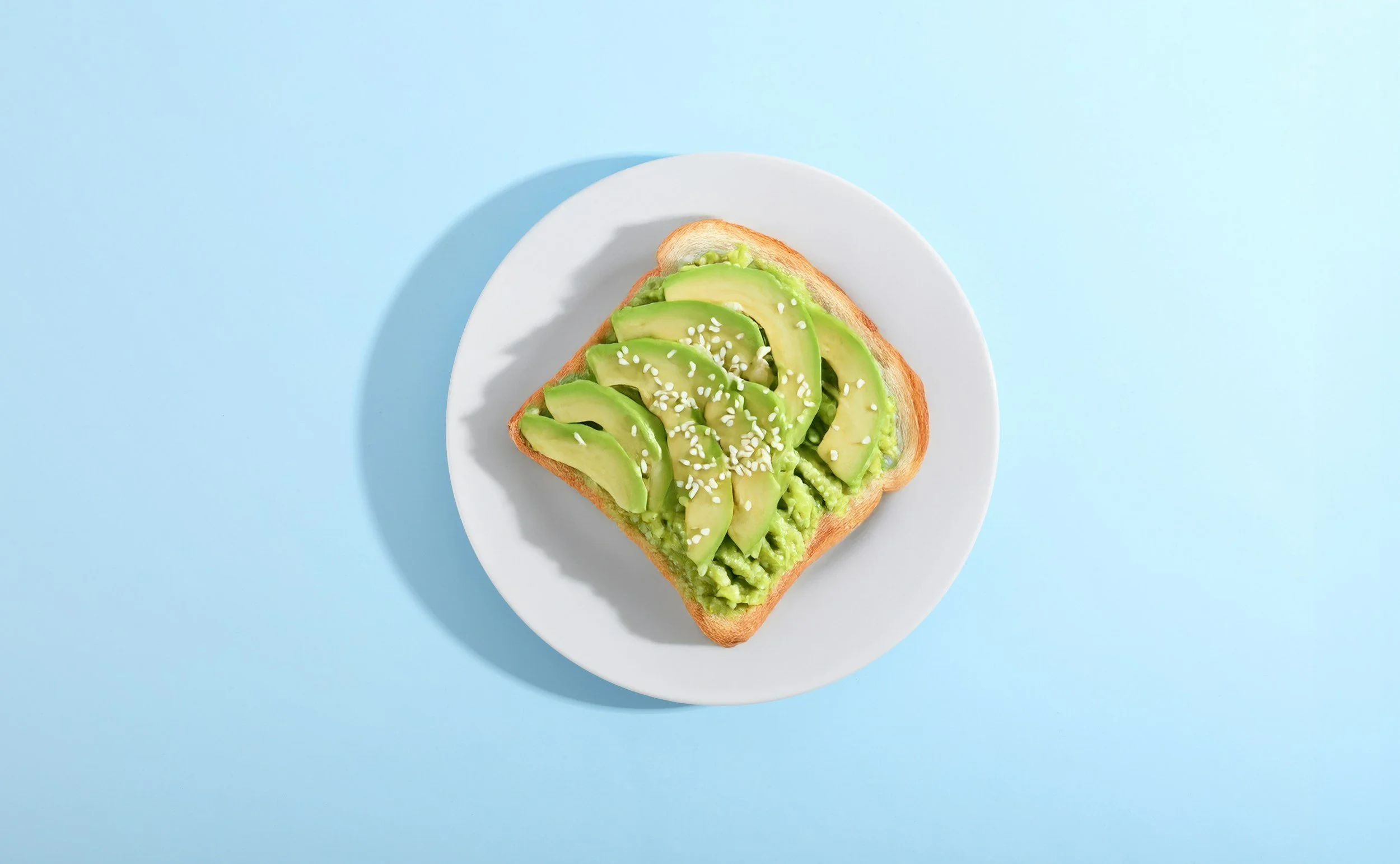 A single piece of toast on a white plate with a blue background. The toast is covered in fresh slices of avocado. There are sesame seeds sprinkled on top.