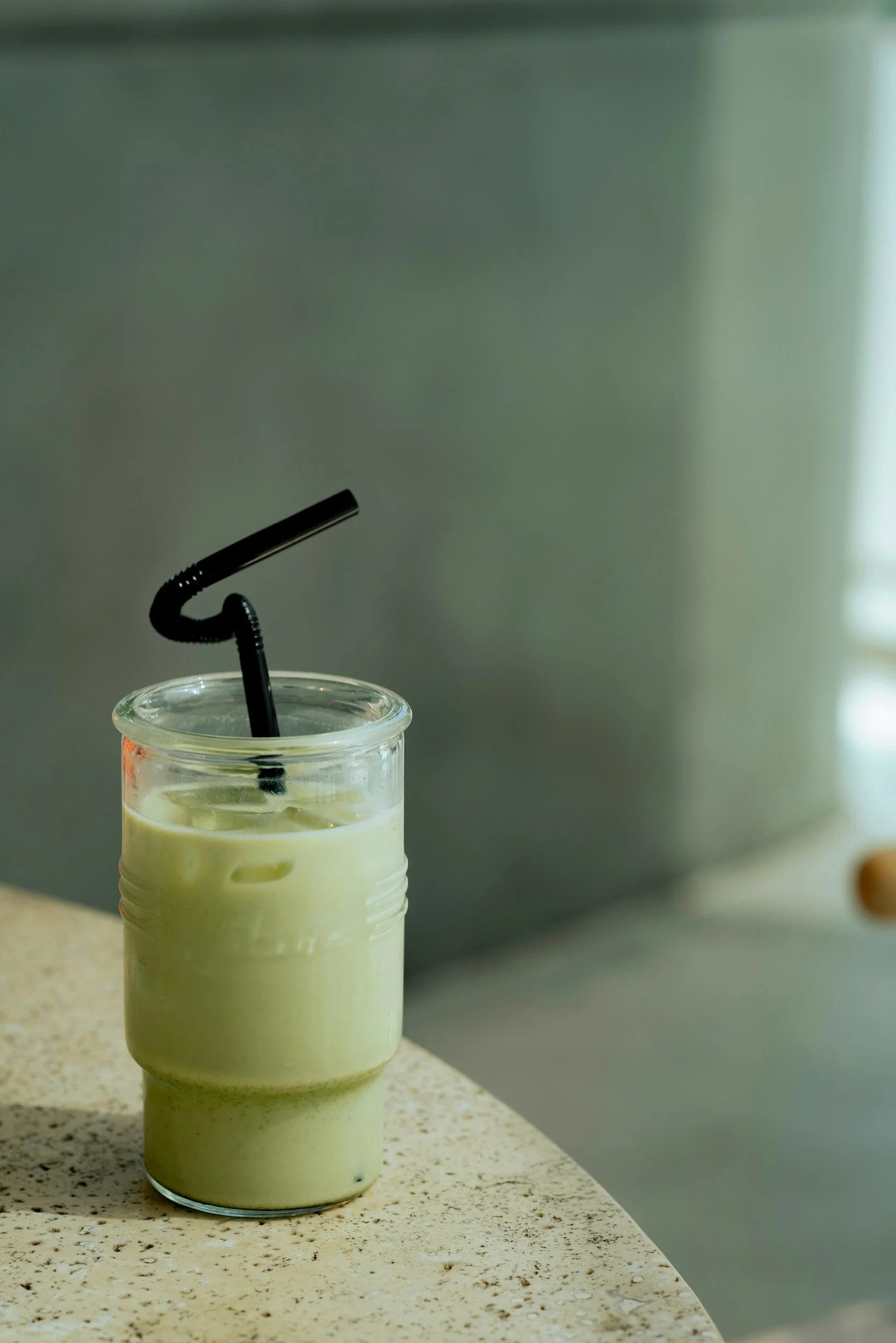 A green avocado smoothie in a glass cup. There is a black twisted straw in the smoothie. The smoothie is sitting on a white marble table top.