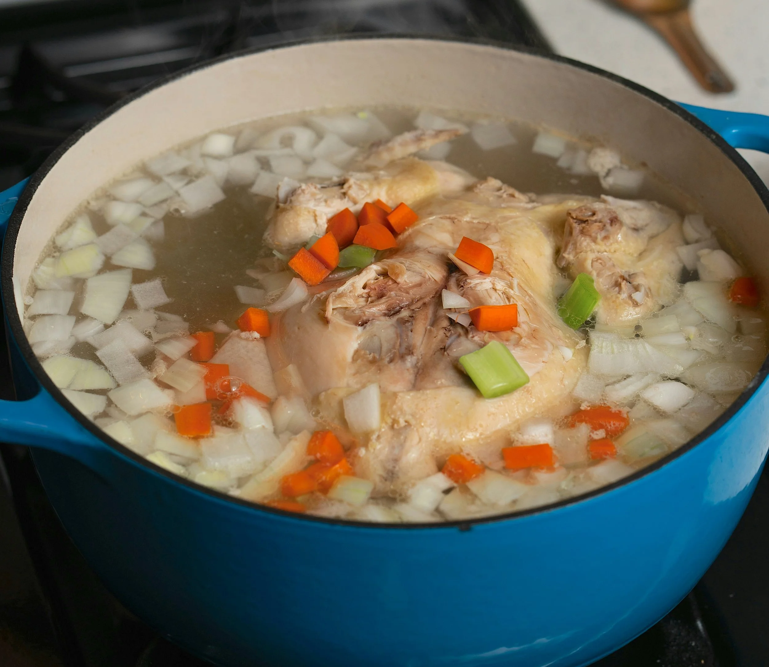 a blue pot with chicken stock cooking inside. There is broth, a full chicken, carrots, celery, and onion inside. The pot is on a black stove.