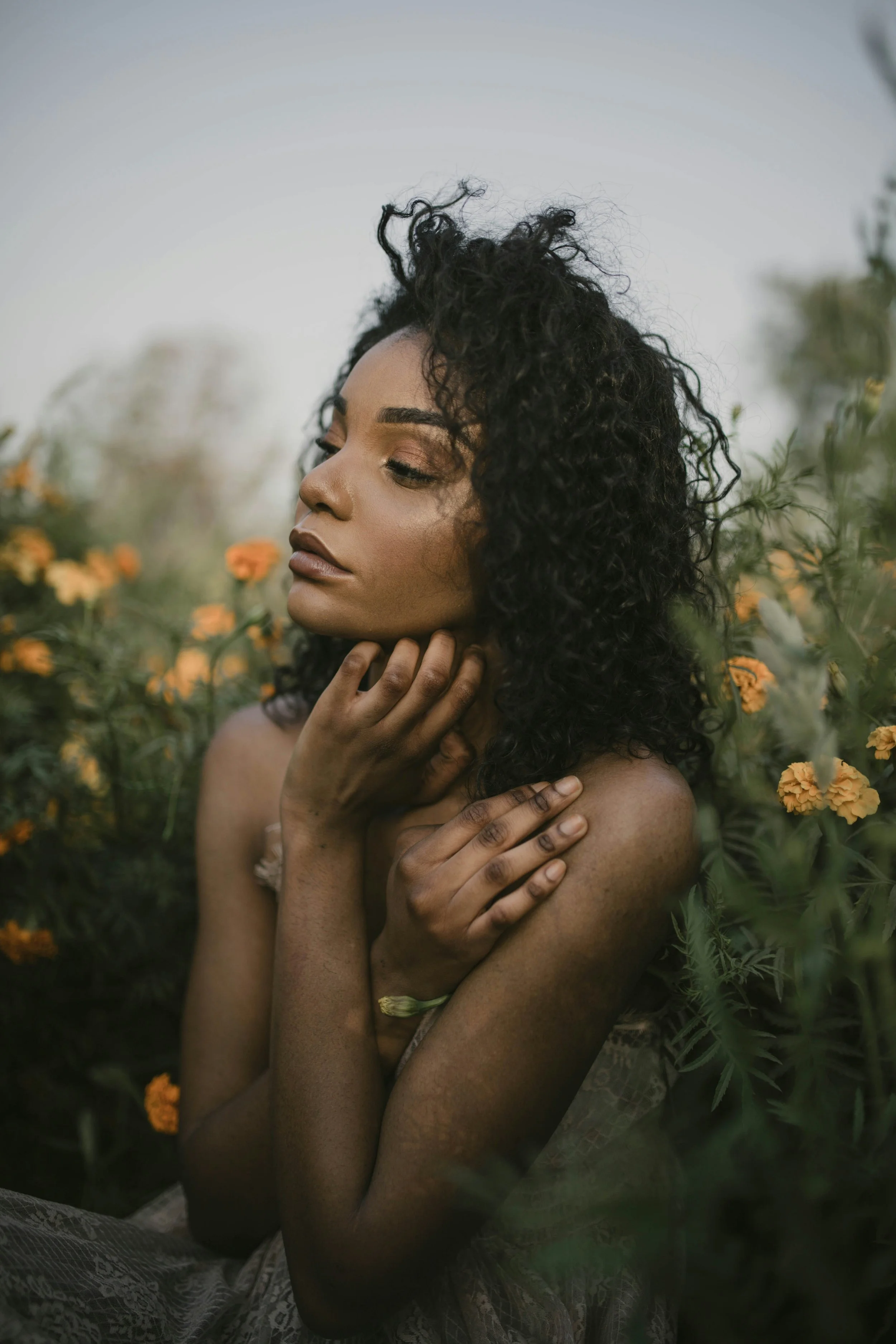 an African American woman gazing into the distance. She has tousled, curly hair. Her hand is resting on her chin. She is sitting in front of a field of wildflowers. She is wearing a billowing off-white dress