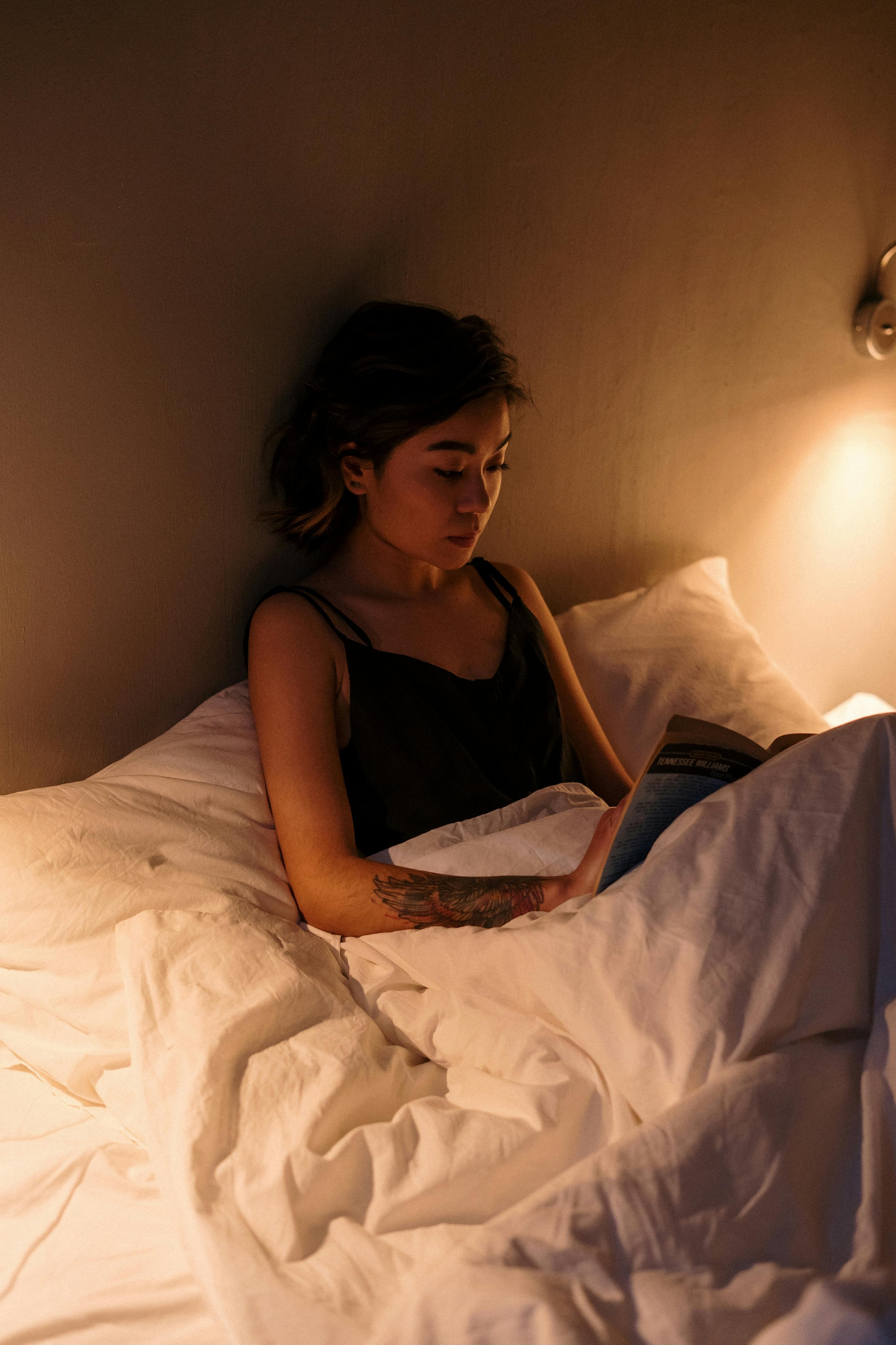 A woman reading in bed with a pale yellow light by her bedside. She's cozy in all-white bedding. She's wearing a black spaghetti strap sleep shirt.