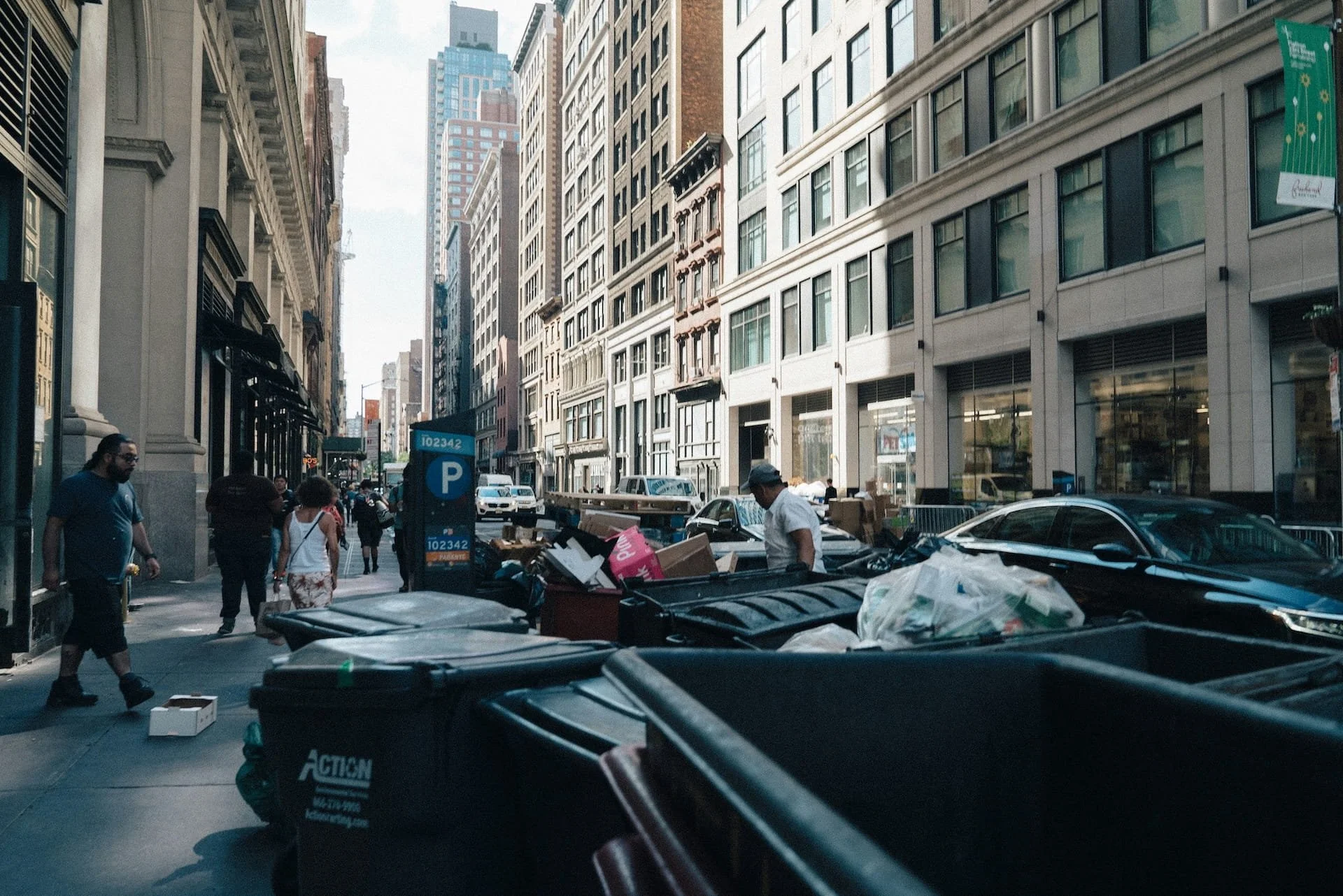 City street with tall buildings, several people walking on sidewalk, and large dumpsters filled with trash and boxes.
