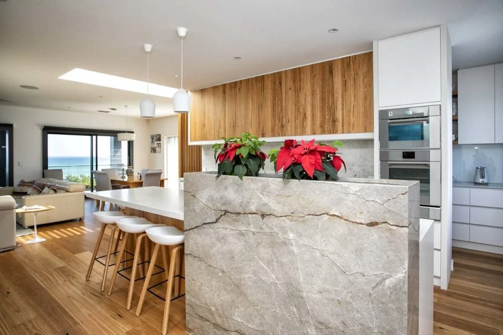Modern open-concept kitchen with white cabinets, black and stainless appliances, and a marble island with red poinsettia plants. Connected to a living room with large windows and ocean view.