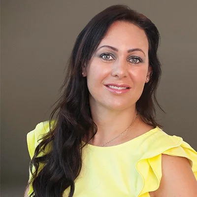A woman with long dark hair and blue eyes, smiling, wearing a yellow top with ruffle sleeves, against a neutral background.
