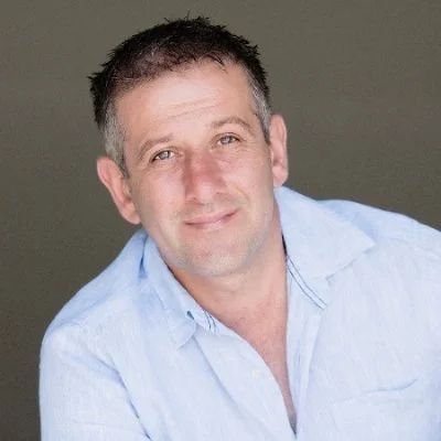 A man with short dark hair and a light beard, wearing a light blue button-up shirt, smiling at the camera against a plain background.