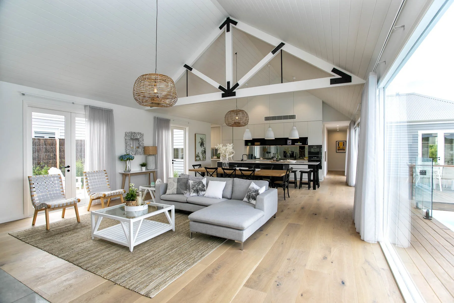 Open-concept living room with white walls, a gray sectional sofa, wooden chairs, and a large sliding glass door leading to an outdoor deck, with a high vaulted ceiling featuring exposed beams and modern lighting fixtures.