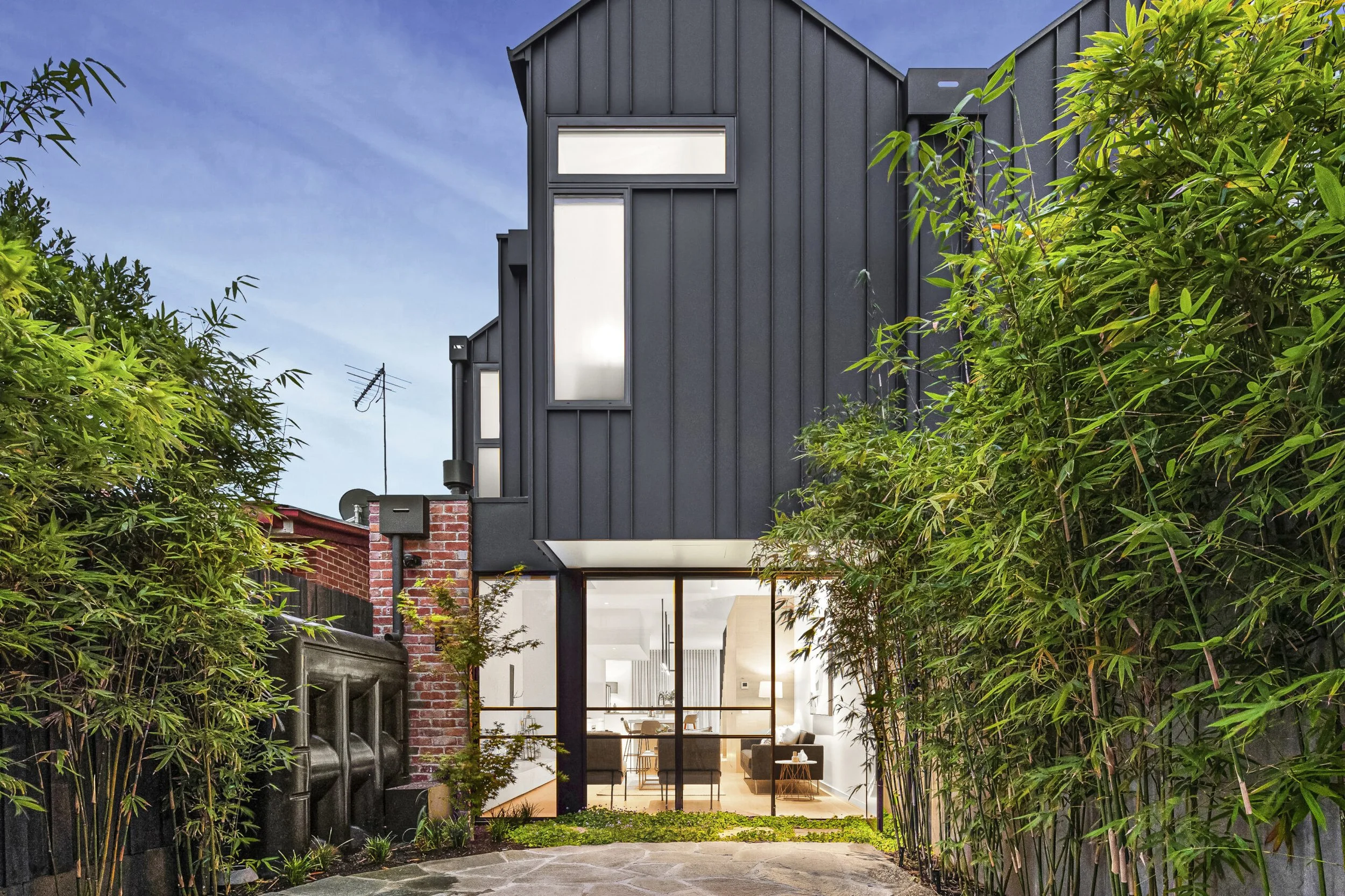 Modern black house with large glass doors and windows, surrounded by lush greenery, during dusk.