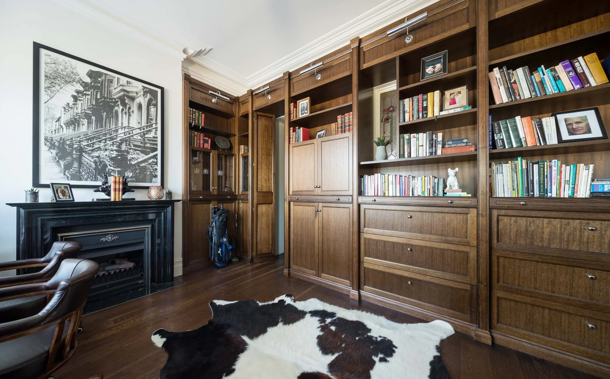 A home library with wooden bookshelves filled with books, a cowhide rug on the wooden floor, a decorative black fireplace, chairs, framed photographs, and artwork on the wall.