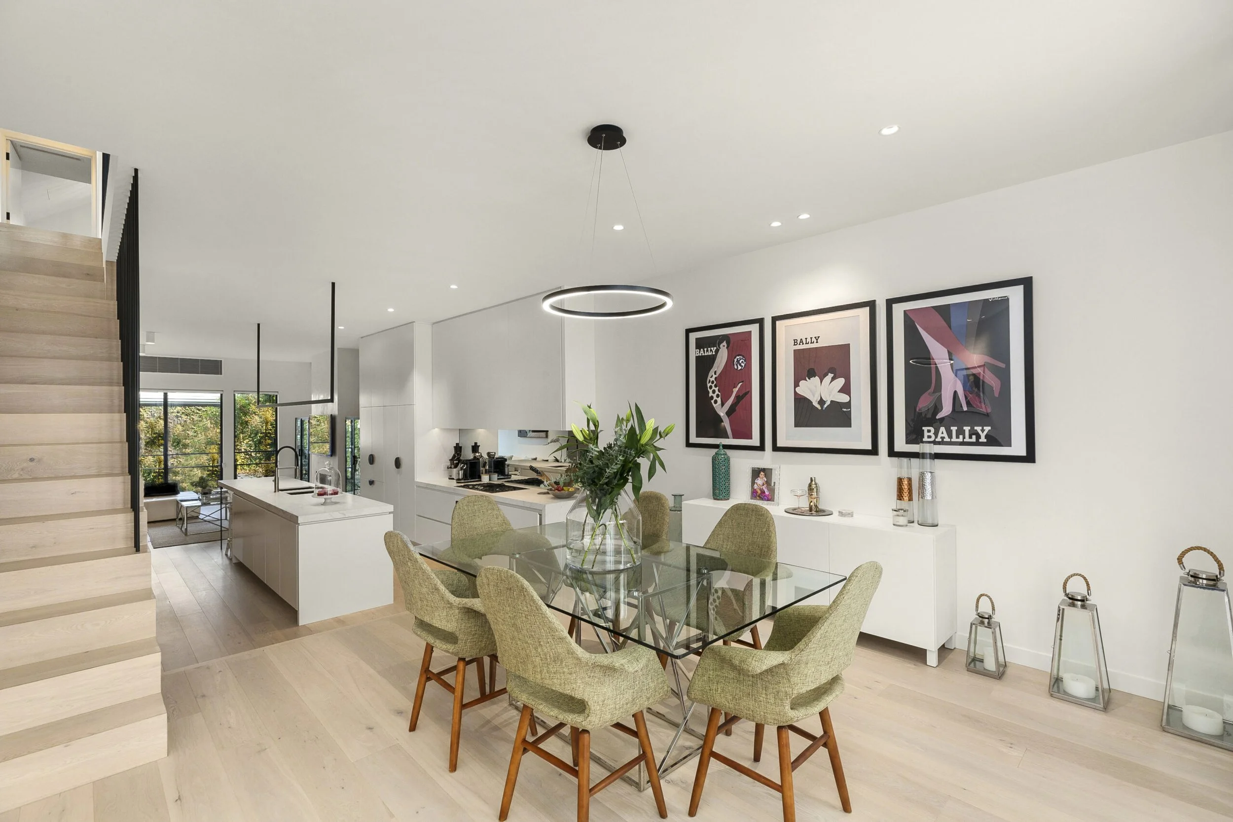 Modern dining area with a glass table, seven green upholstered chairs, and a white sideboard with framed art pictures on the wall. The space has a neutral color scheme with light wood floors and a staircase to the left.