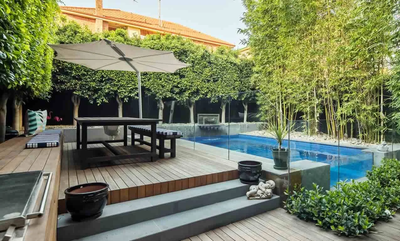 Outdoor backyard with a swimming pool, wooden deck, patio table with an umbrella, cushioned seating area, potted plants, and surrounded by lush green trees and bamboo.