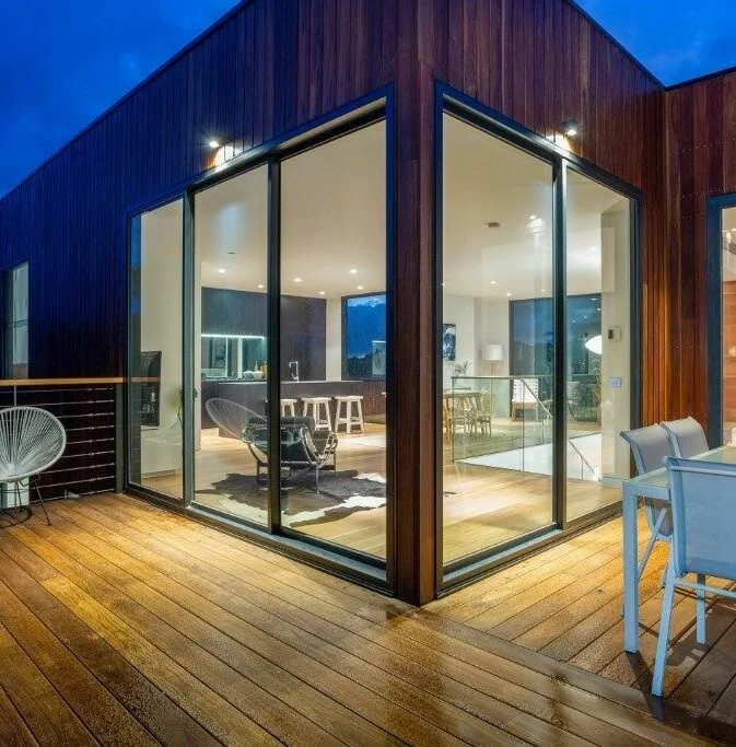A modern house with large glass windows and sliding doors, wooden exterior, and a wooden patio with outdoor chairs and furniture, illuminated in the evening.