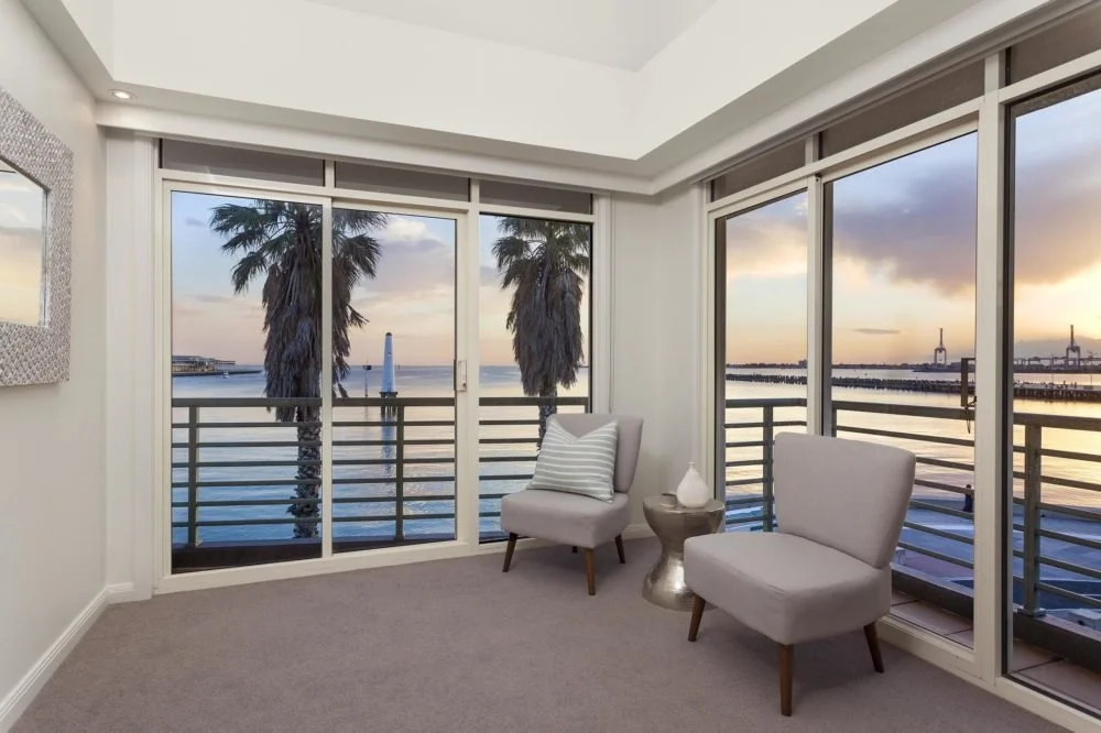 Living room with large glass windows and sliding doors overlooking a waterfront with two palm trees, railings, and a sunset sky. Two gray chairs, a small metallic side table, and a striped pillow.