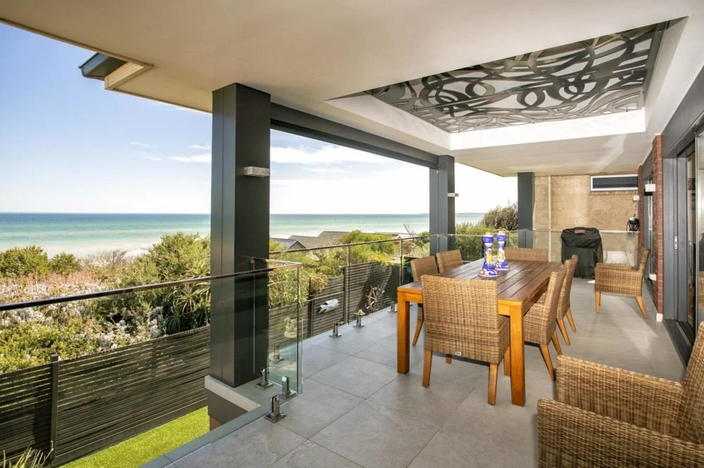 A spacious balcony with a dining table and chairs overlooking a beach and ocean under a partly cloudy sky.