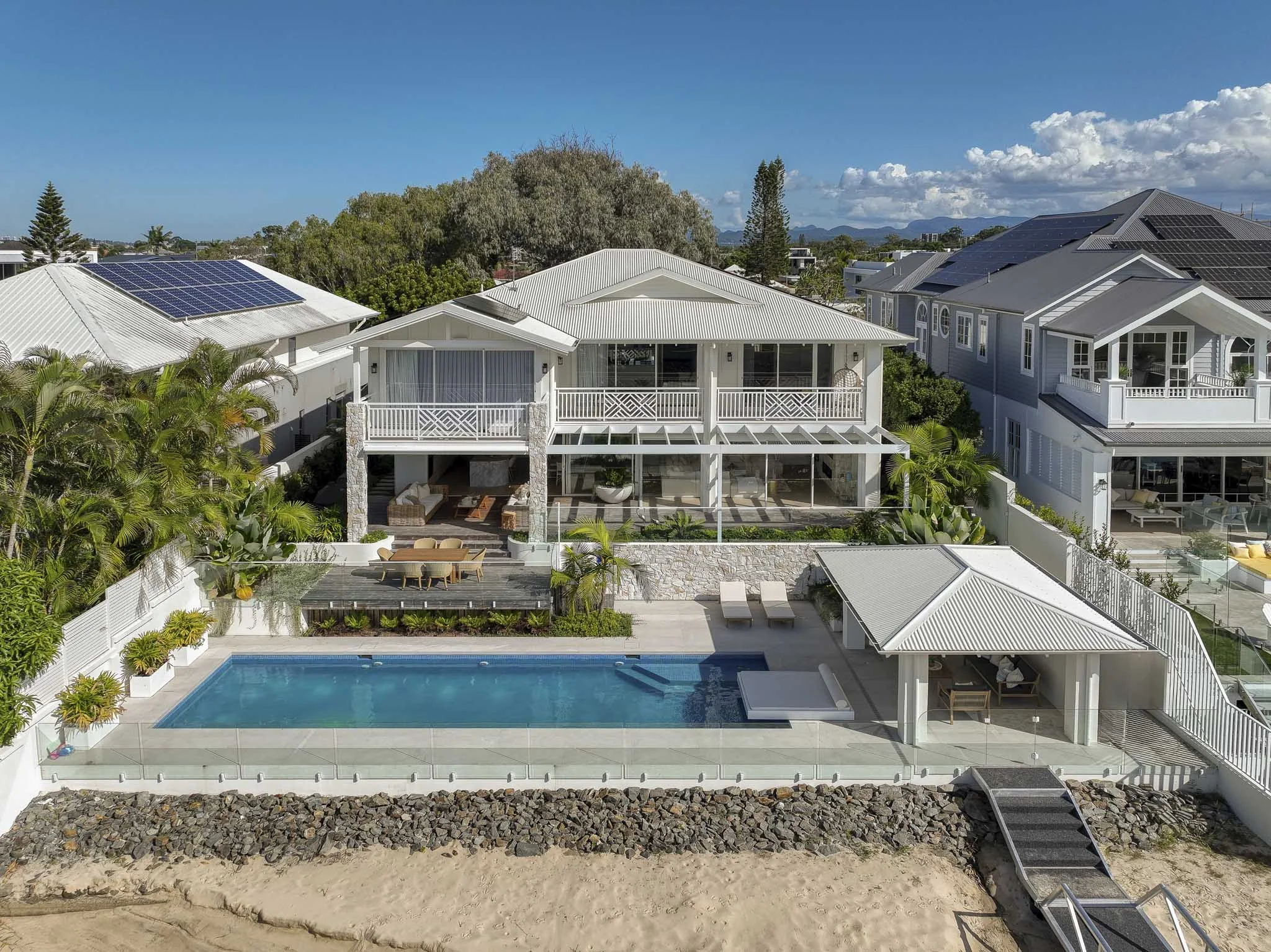 Aerial view of a modern house with a swimming pool in backyard, surrounded by lush greenery and neighboring houses with solar panels on roofs.