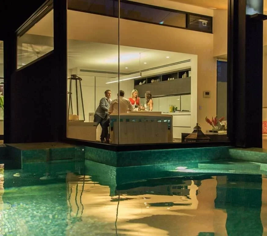 People gathered in a modern kitchen seen through a glass wall, with a pool in the foreground at night.