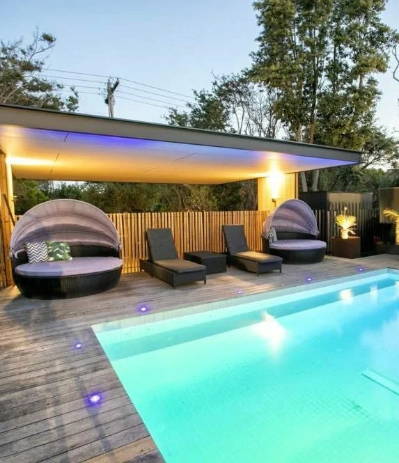 Modern backyard swimming pool area with lounge chairs and round sunbeds under a shaded patio at sunset.