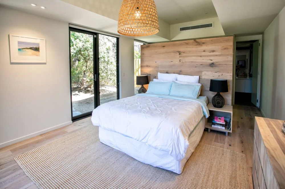 Bedroom with a bed, two black lamps, wooden headboard, sliding glass door, woven light fixture, and a view of greenery outside.