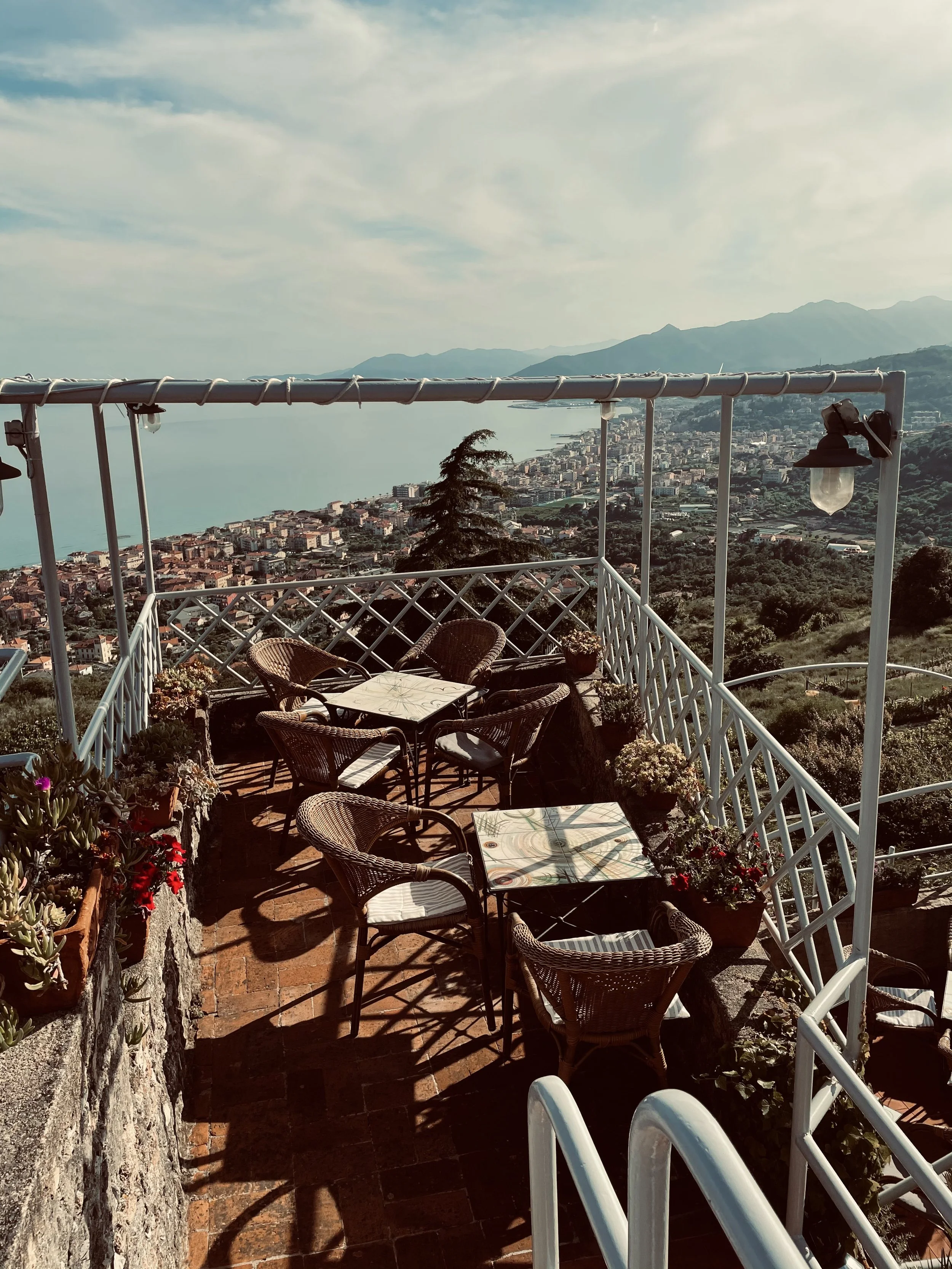 Terraza con tavoli e sedie di vimini, fiori in vasi, vista panoramica sulla costa e sui monti al tramonto.
