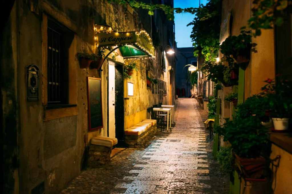 Una strada stretta e pittoresca con edifici di pietra e piante, illuminata da luci calde, che sembra una via di un villaggio italiano, di sera.