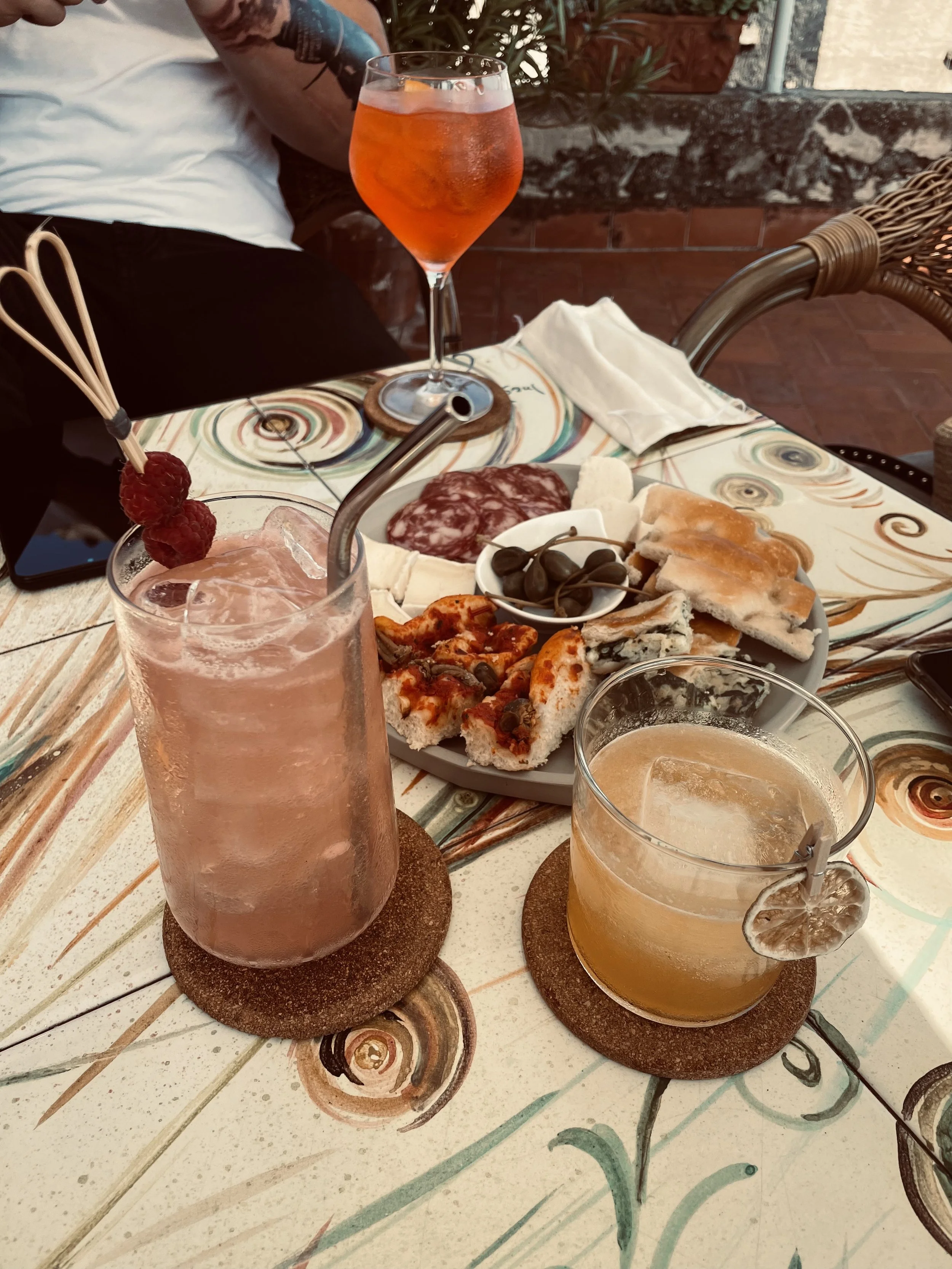 Una tavola apparecchiata con due cocktail, uno rosa con lamponi e uno giallo con una fetta di limone, e un piatto di antipasti con salumi, formaggi, focaccia e olive in un ambiente esterno.