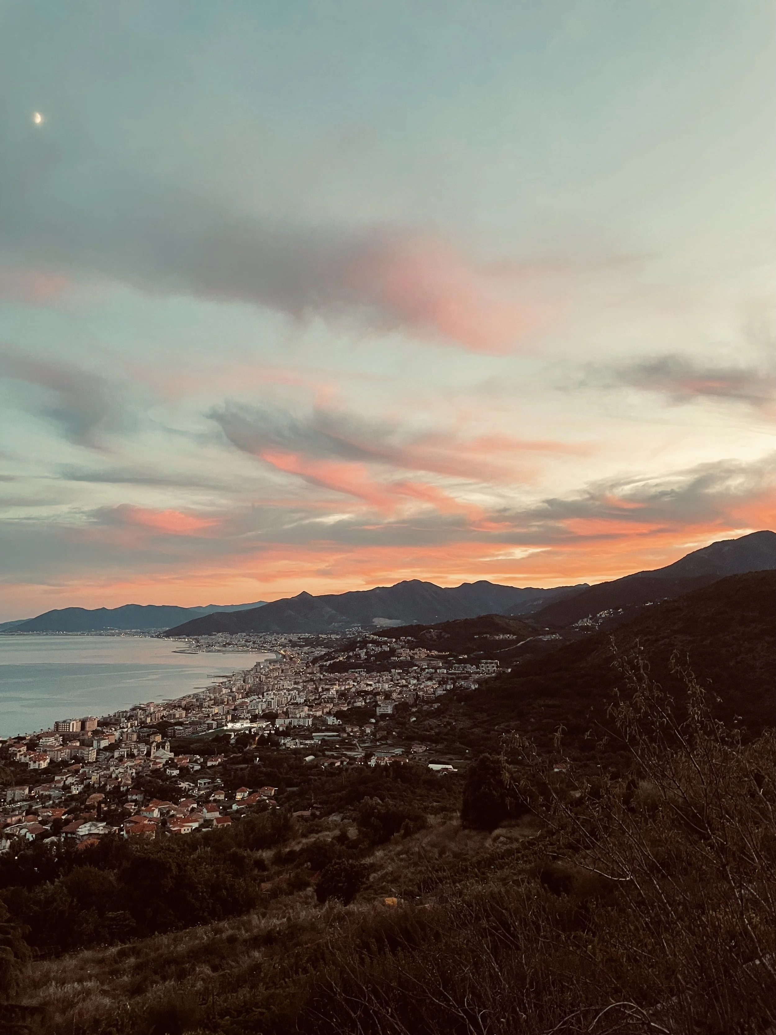 Paesaggio al tramonto con mare, città costiera, montagne e cielo con nuvole rosa e blu