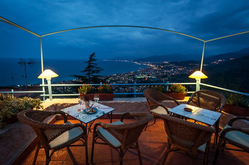 Terrazza con tavolo e sedie, luci da tavolo, vista panoramica sulla costa e città di notte.