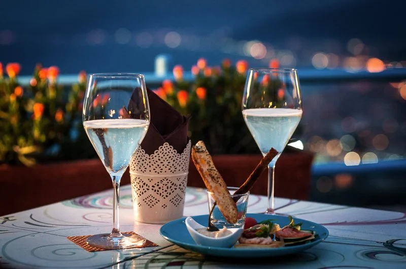Due calici di vino bianco su un tavolo, con un piatto di aperitivo e un'illuminazione soffusa, sfondo di città illuminata di notte.