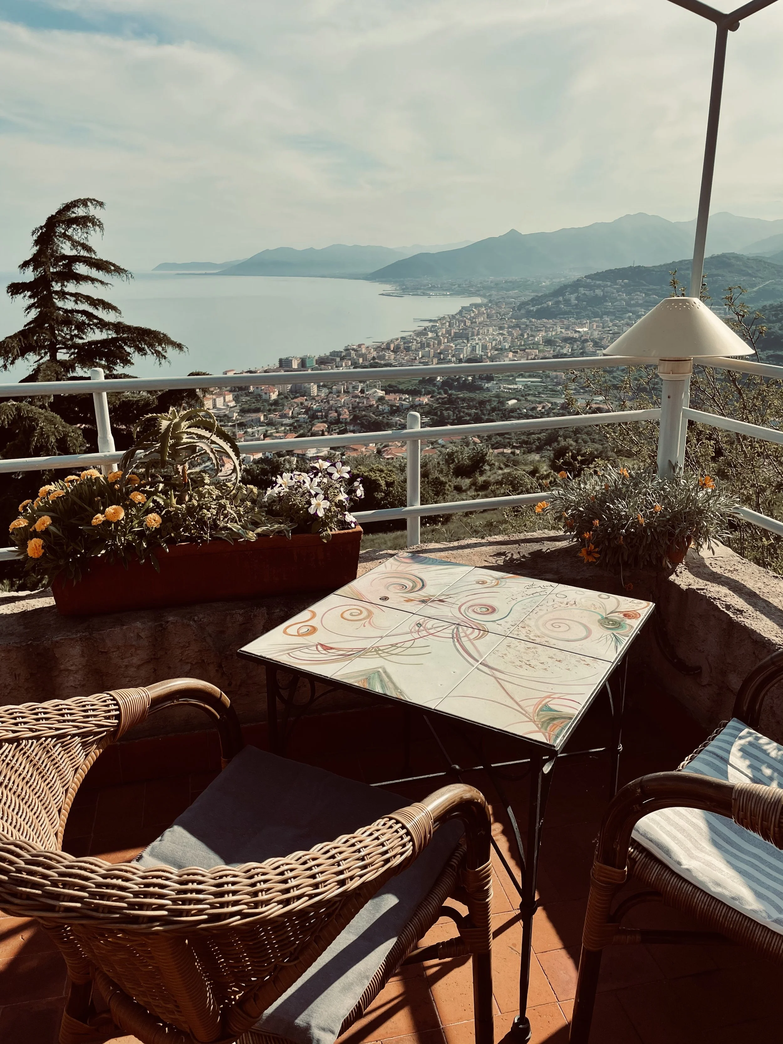 Vista panoramica da un terrazzo con tavolino decorato, sedie in vimini, fiori colorati su una pianta, e una vista sul mare, le montagne e la città sottostante