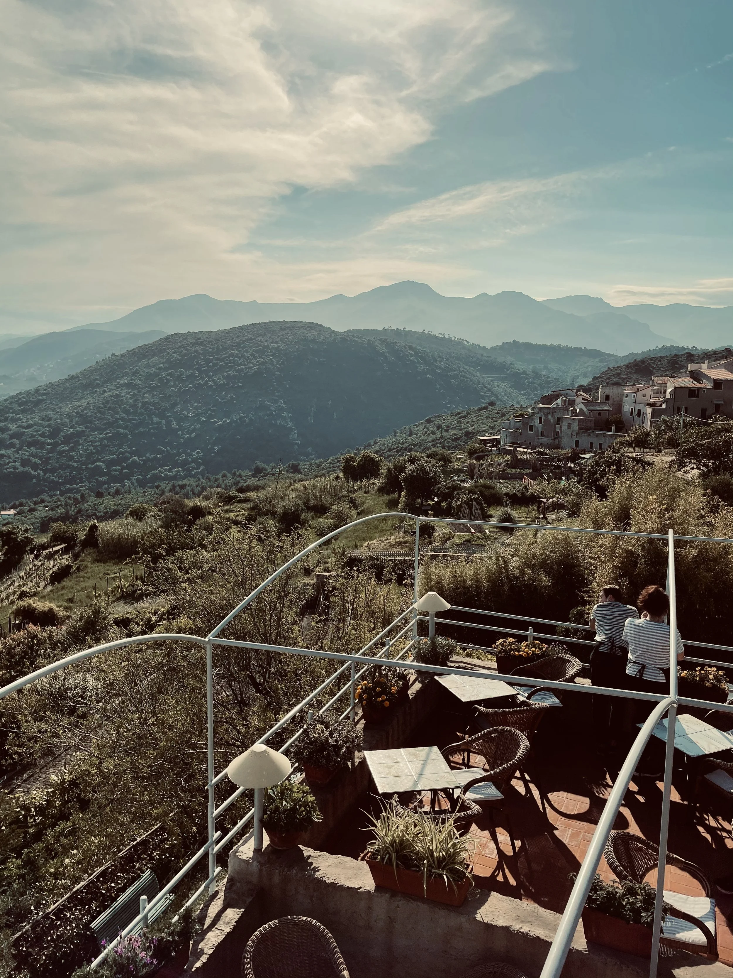 Vista di un terrazzo con tavoli, sedie e piante, che guarda verso le colline e le montagne in lontananza, con persone sedute e che chiacchierano.