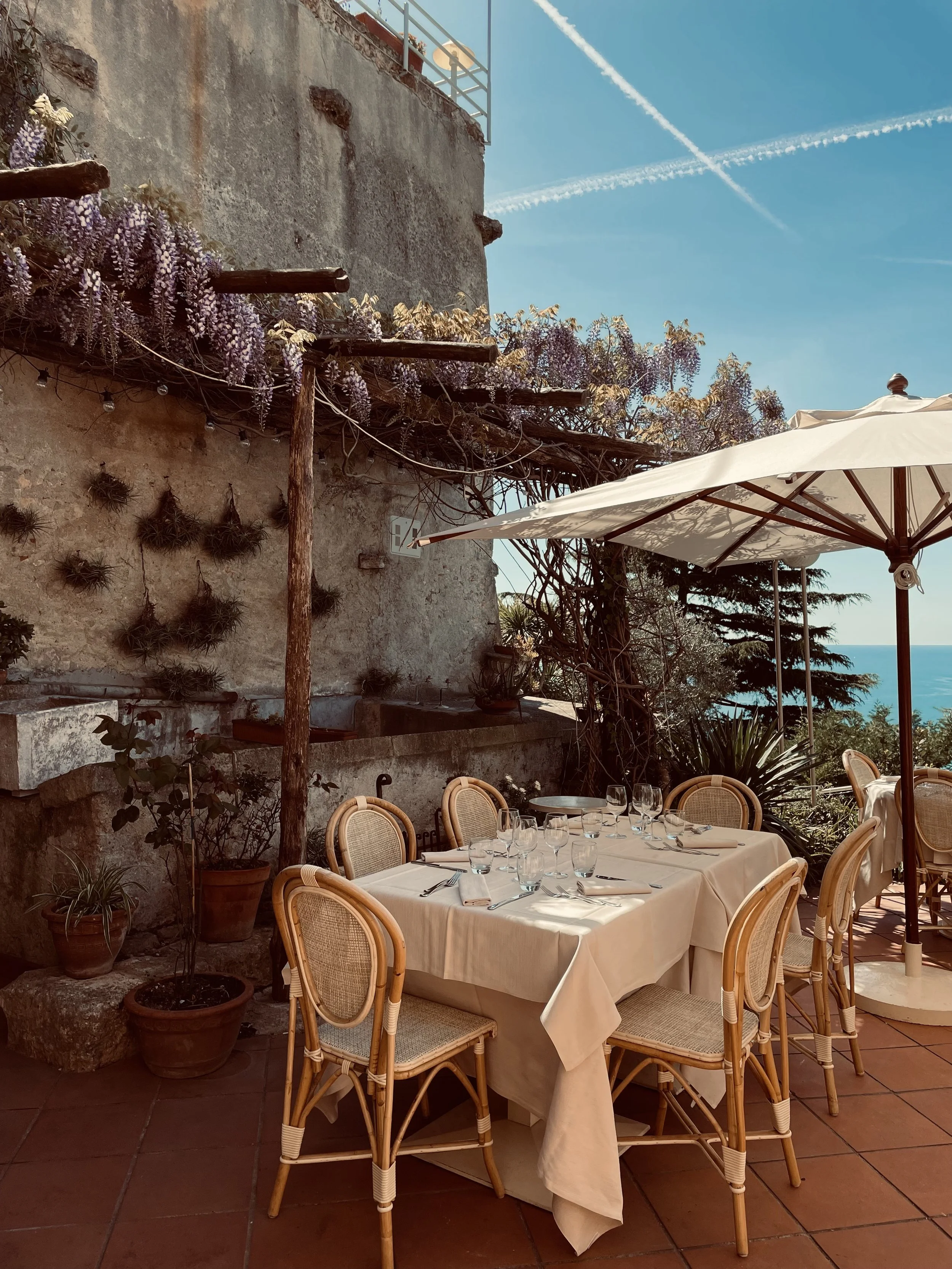 Tavolo apparecchiato con tovaglia bianca, bicchieri e posate, in un ristorante all'aperto con sedie di vimini, ombrellone e piante in vaso. Sfondo di mura antiche e vista sul mare.