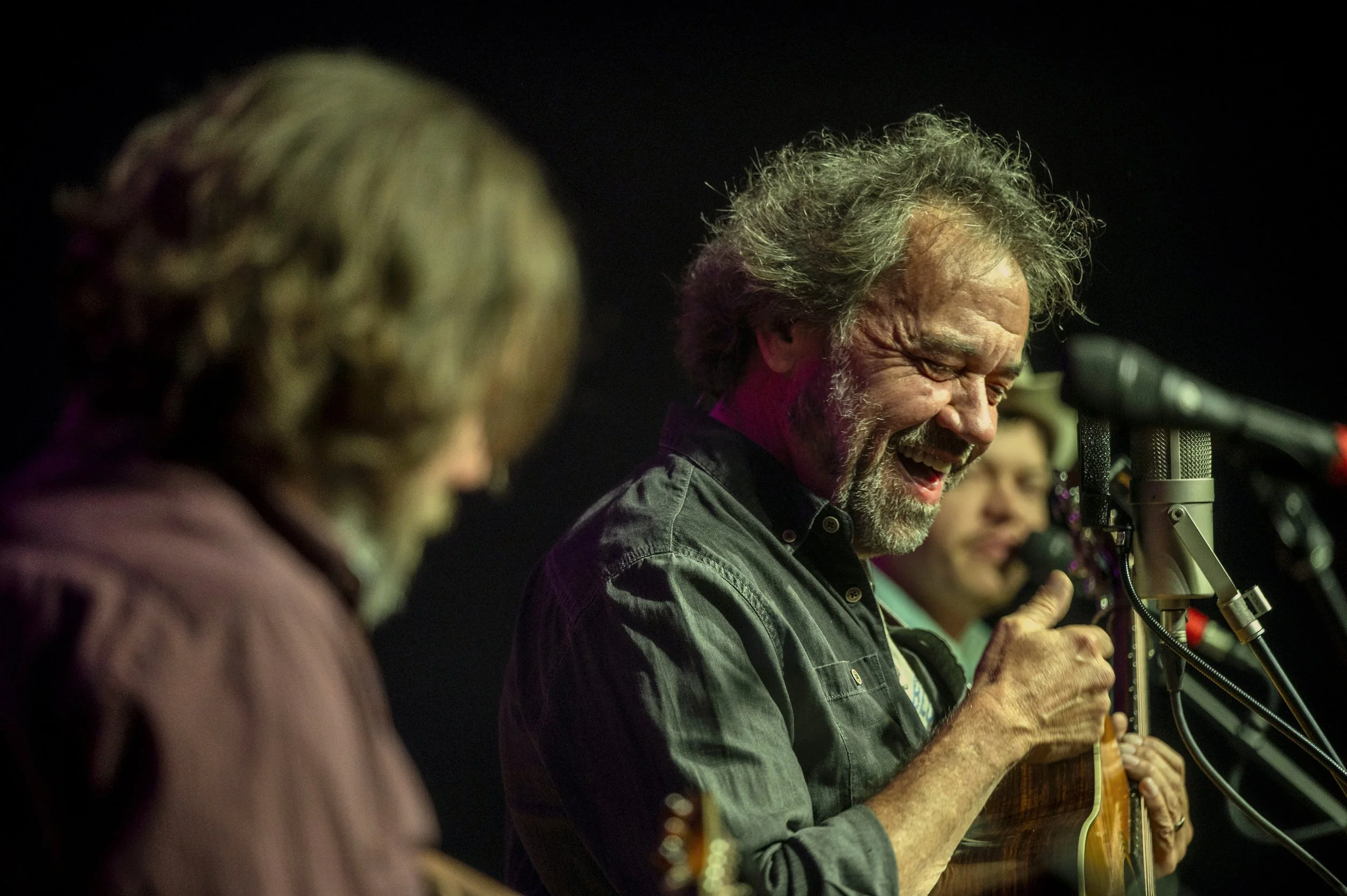 Musicians performing on stage, smiling and playing guitars, with microphones, under dim lighting.