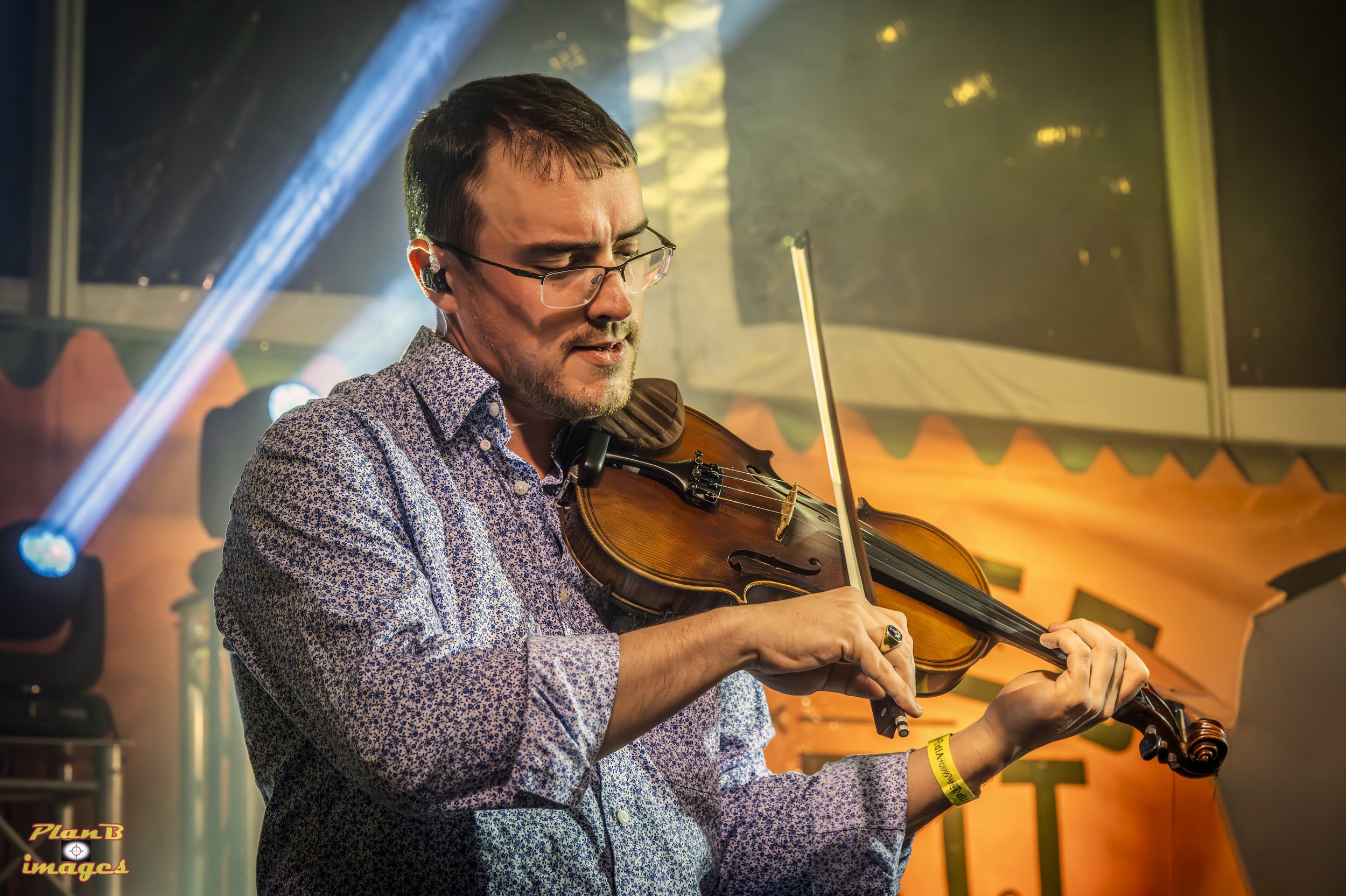 A man with dark hair, glasses, and a patterned shirt playing a violin inside a colorful, festive setting with yellow and green decorations and a plastic bubble ceiling.