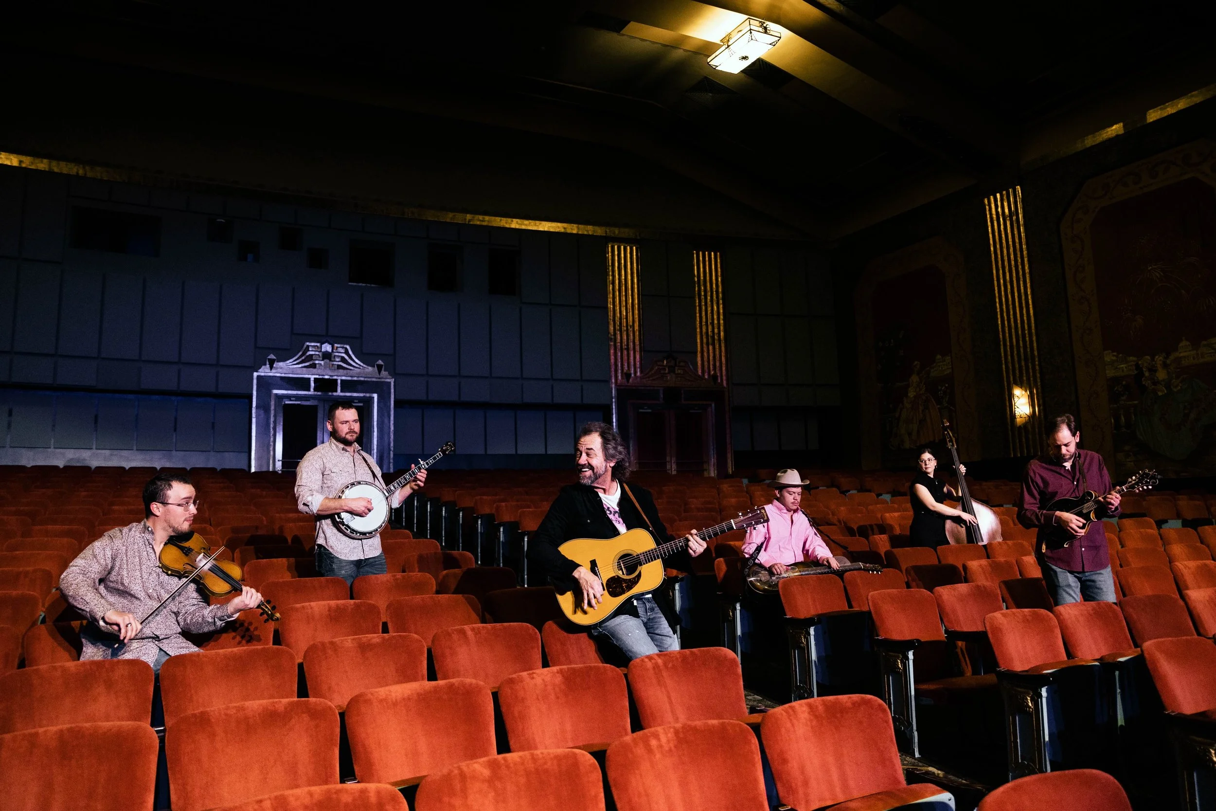 A small musical band practicing in an empty theater with red seats, featuring six members with various instruments including guitars, violin, banjo, double bass, and percussion.